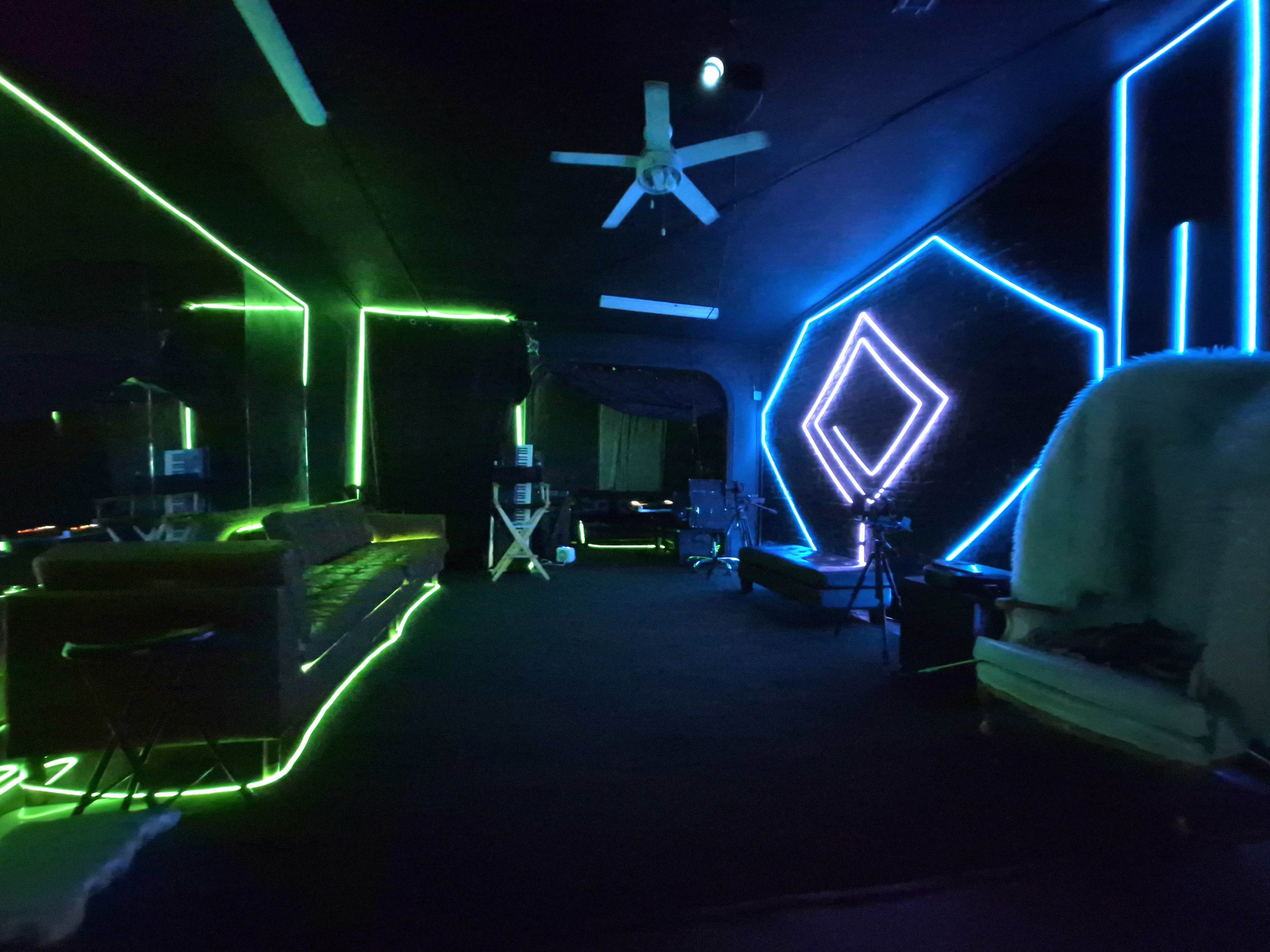The image shows a dimly lit room featuring neon green and blue lights outlined on the walls and furniture.