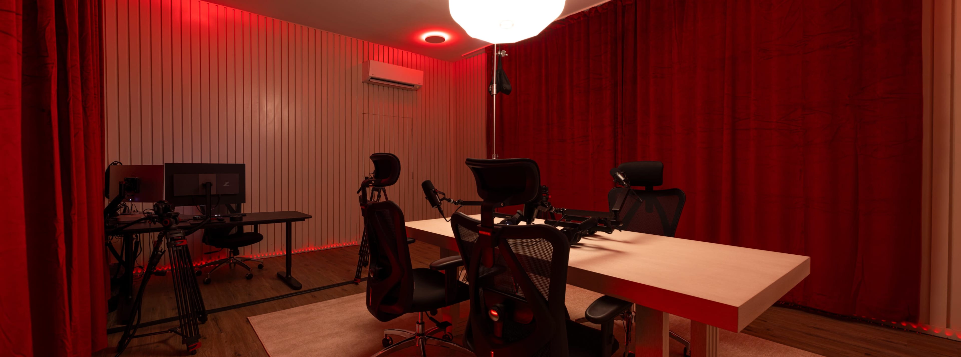 A recording studio with a desk, four chairs, two computer monitors, and red curtains illuminated by a ceiling light.
