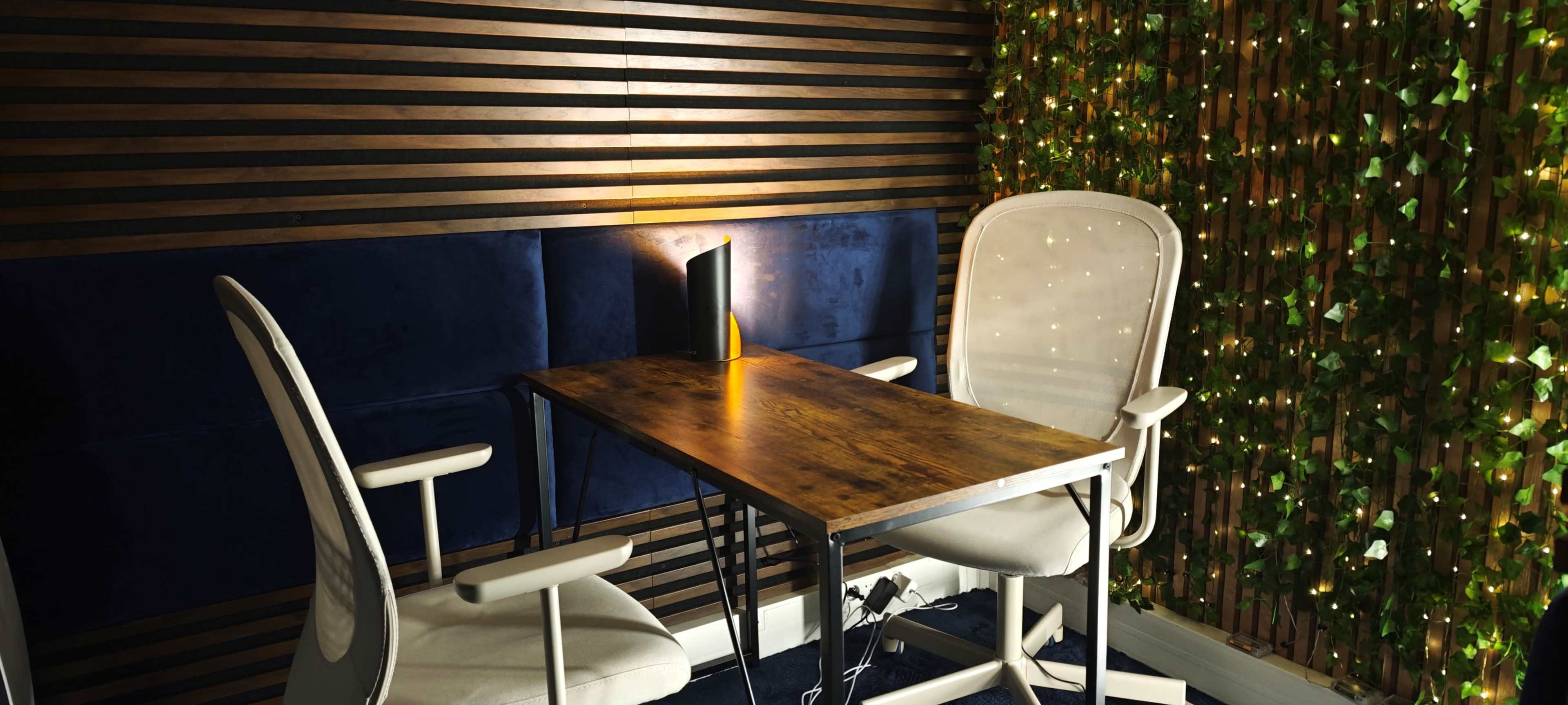 A small meeting space features a wooden table with two white chairs illuminated by a warm light against a backdrop of wood stripes and a wall adorned with greenery.