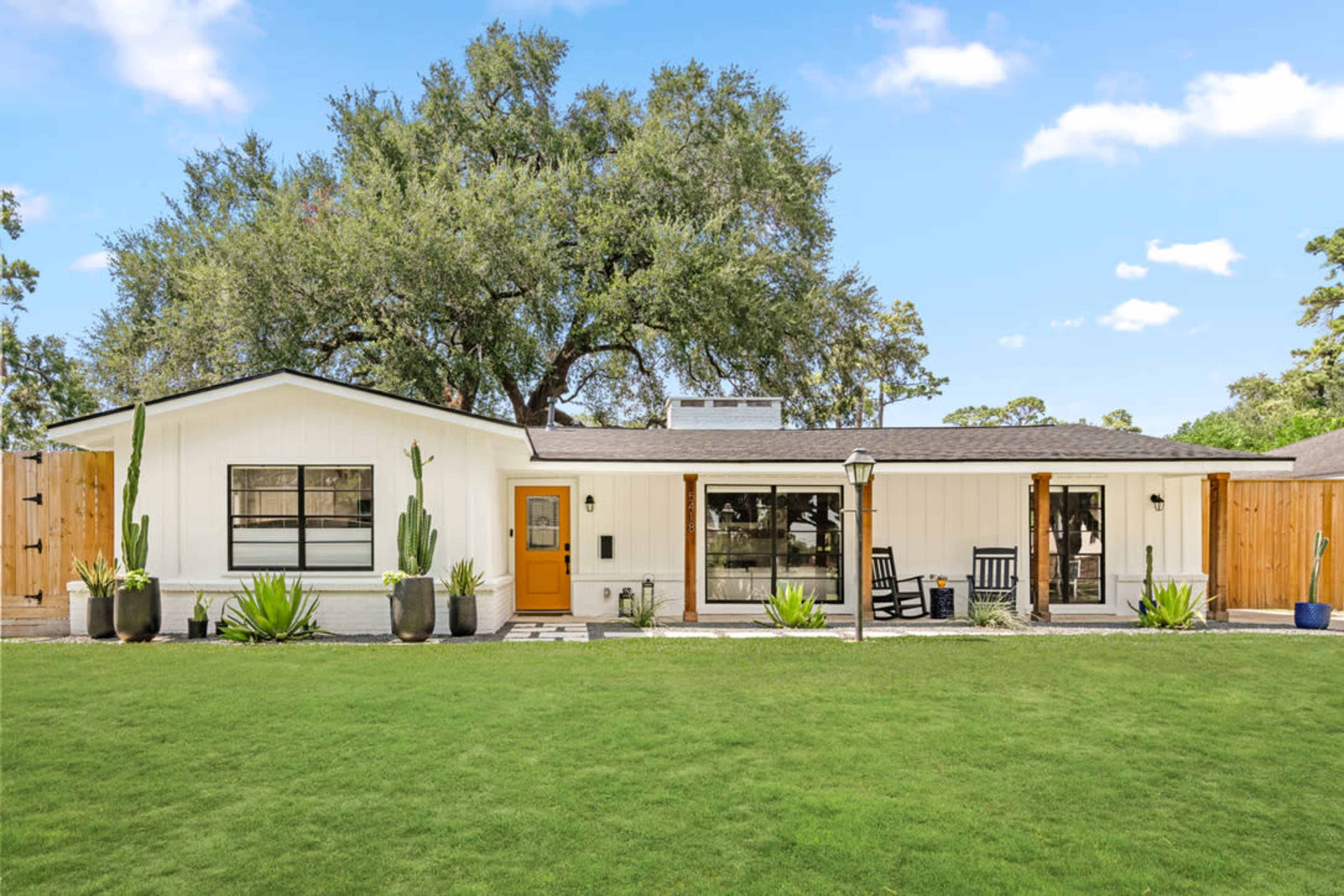 The image shows a single-story house with a light-colored exterior, large windows, a wooden front door, and a well-manicured lawn featuring potted plants and a rocking chair porch.