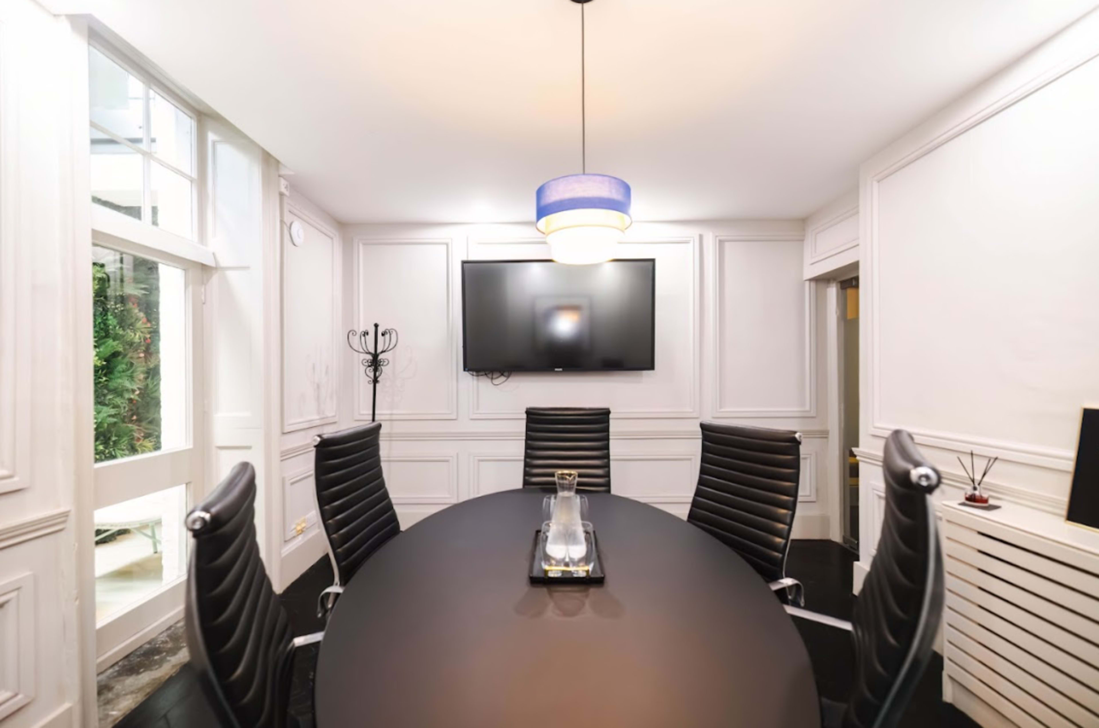 A modern conference room features a large oval table surrounded by black leather chairs, with a television mounted on a white paneled wall.