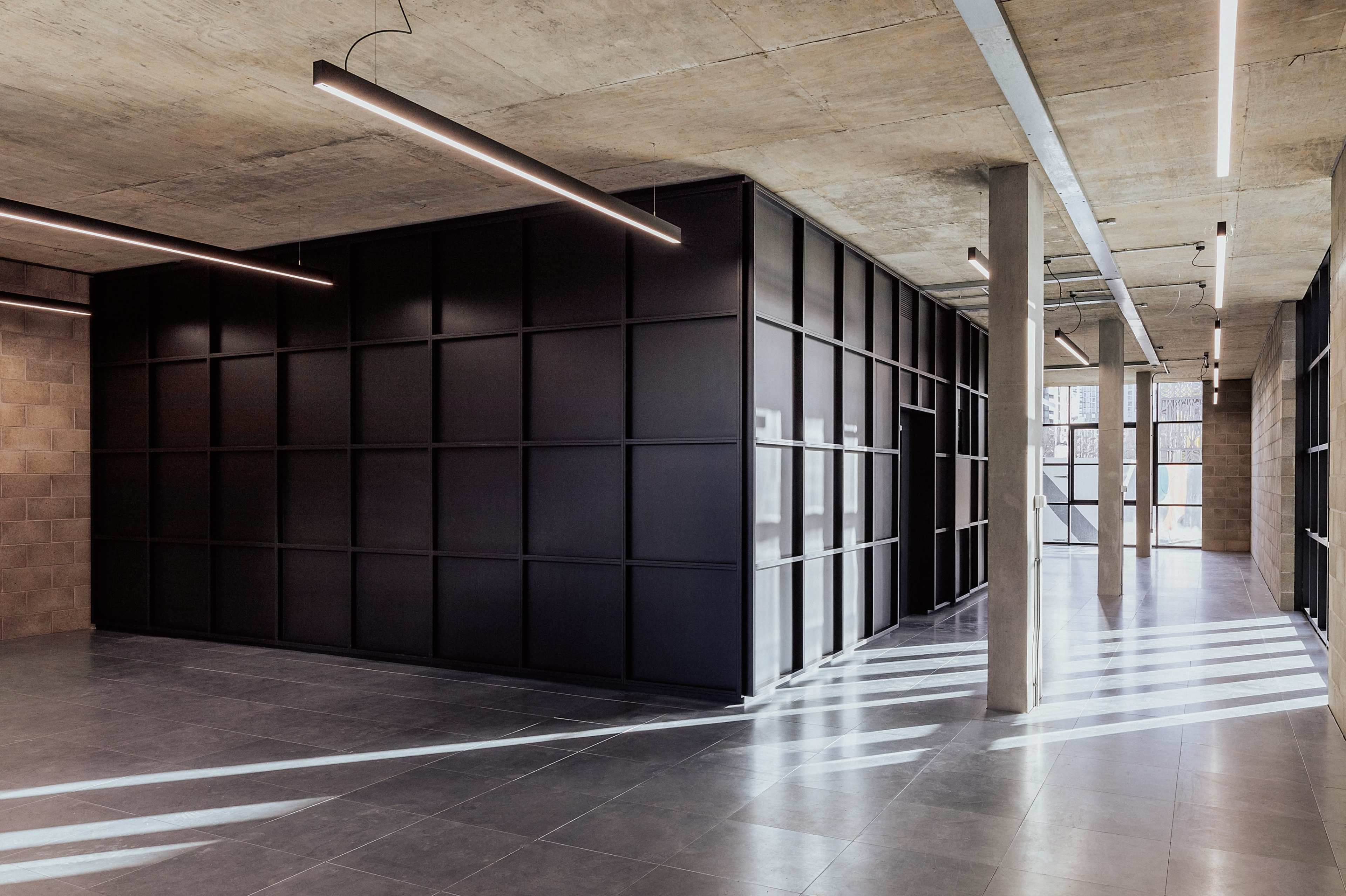 The image shows a modern interior with a polished concrete ceiling, black paneled walls, and large windows casting linear shadows across the floor.