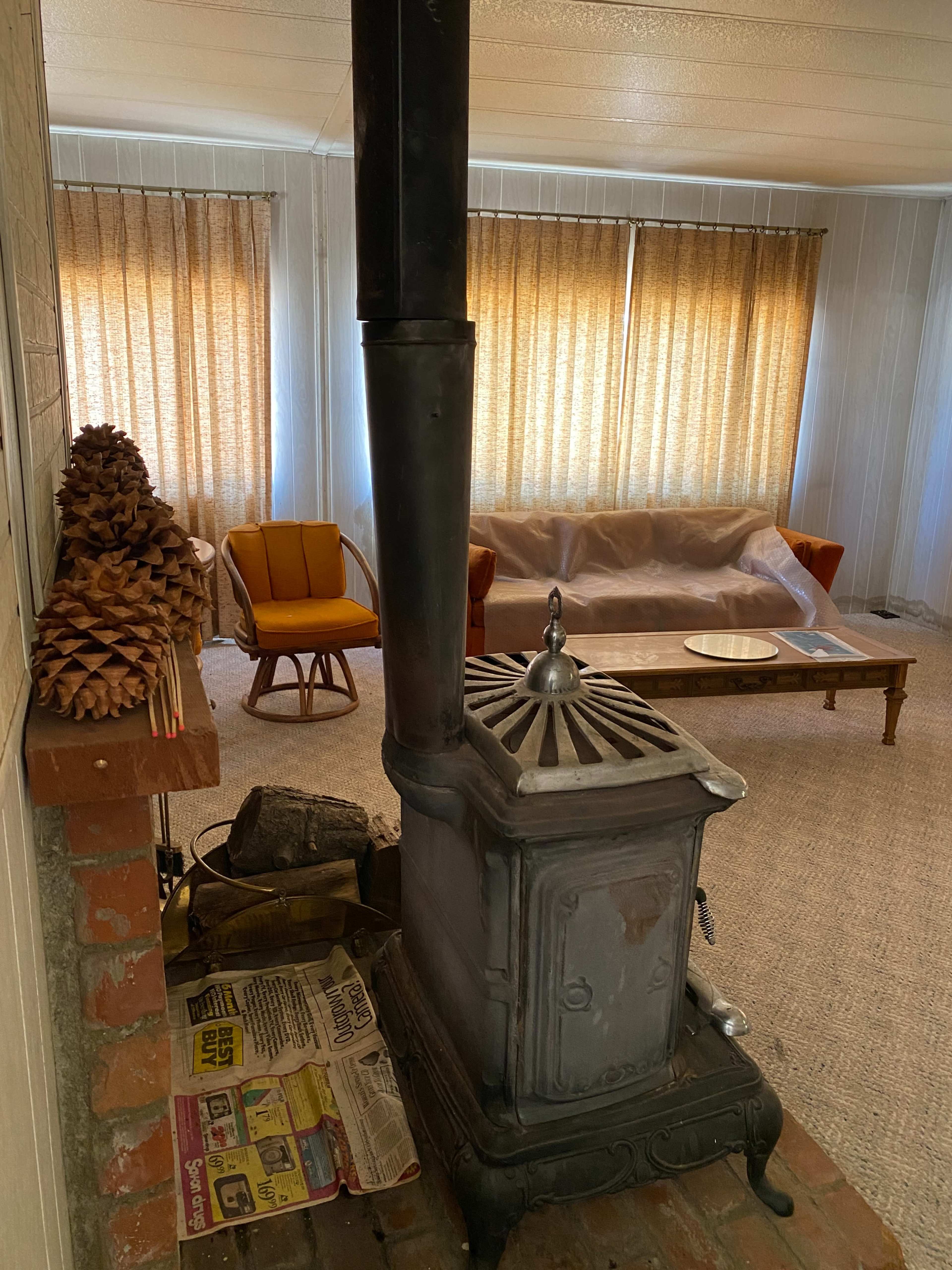 The image shows a cozy living room featuring a wood stove, a wooden coffee table, an orange armchair, and large windows covered with sheer curtains.