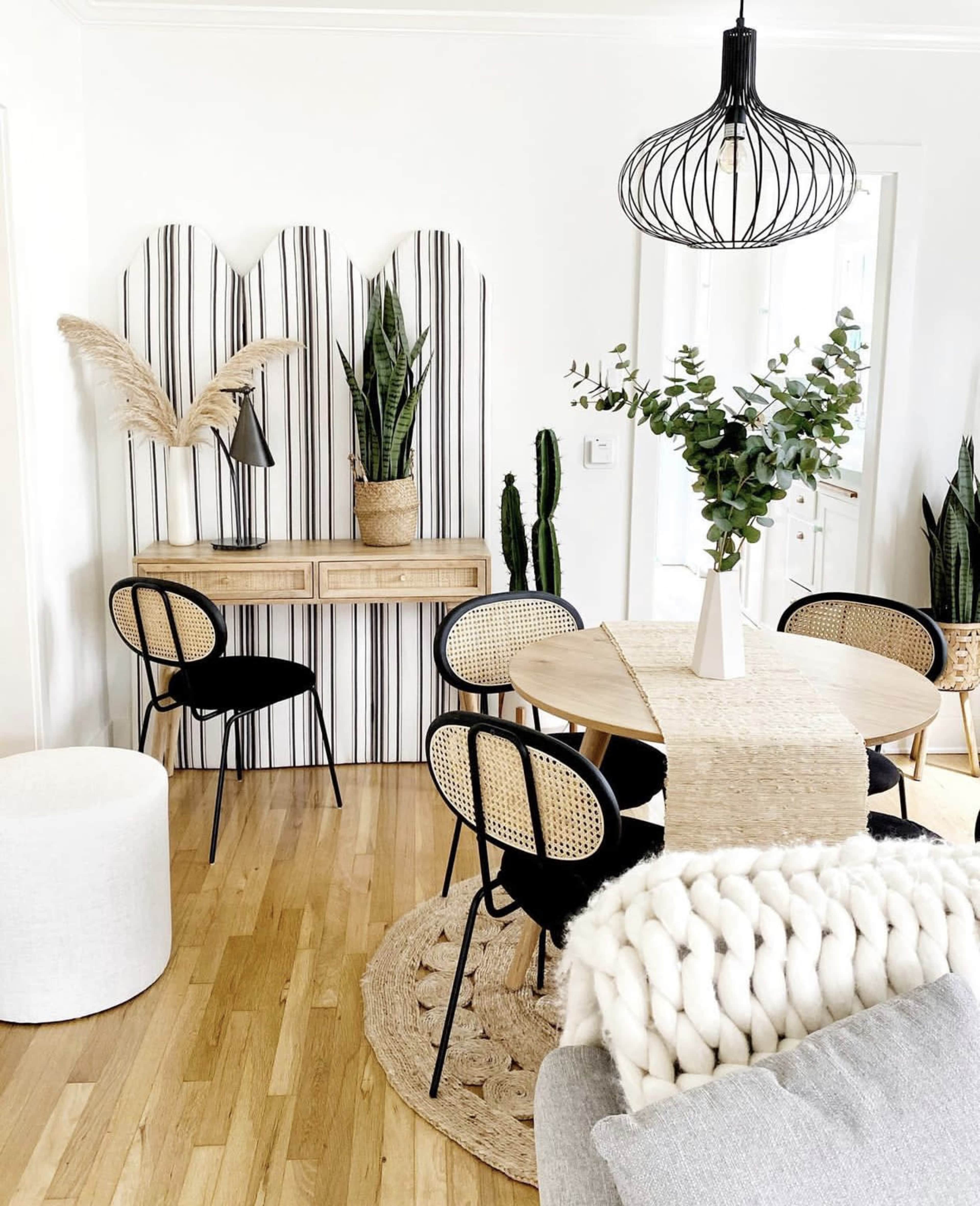 A dining area features a round table surrounded by black and rattan chairs, with a light fixture overhead and various indoor plants in decorative pots.