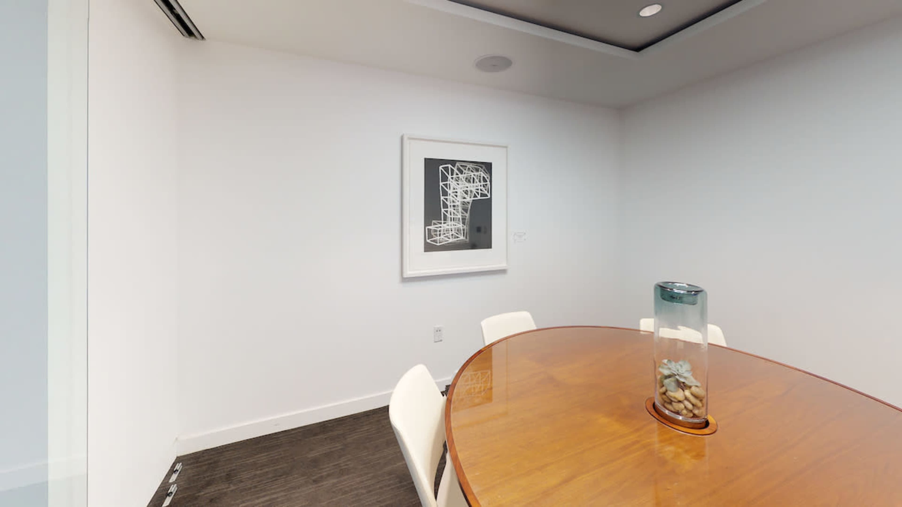 A small, modern meeting room featuring a round wooden table surrounded by white chairs and a framed artwork on the wall.
