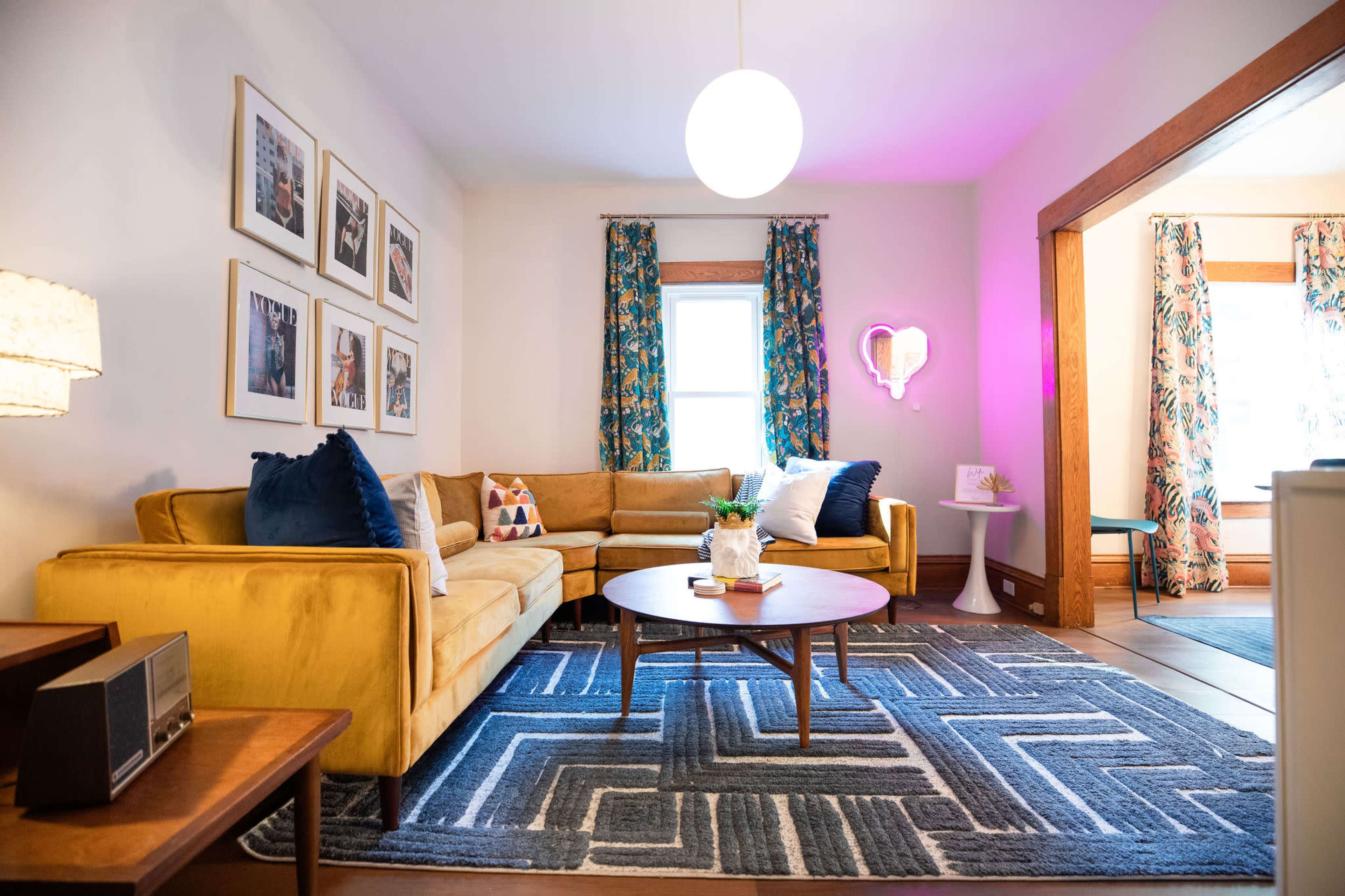 A cozy living room featuring a yellow sectional sofa, a round coffee table, and framed artwork on the walls, complemented by colorful curtains and a stylish area rug.