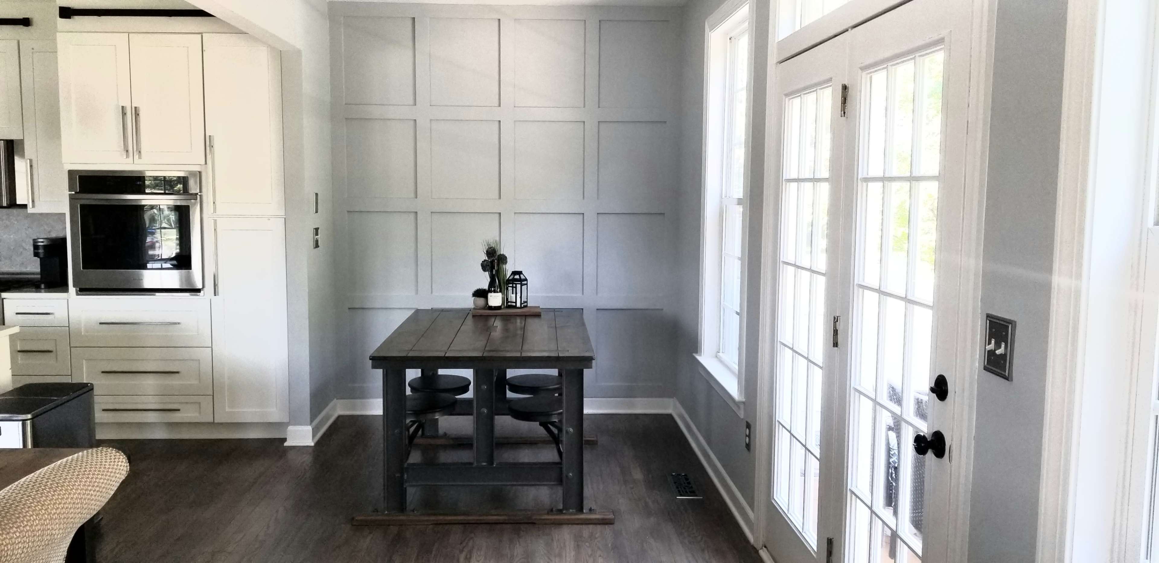 The image shows a dining area with a wooden table and two chairs, adjacent to a wall featuring a grid panel design and large windows with a door leading outside.