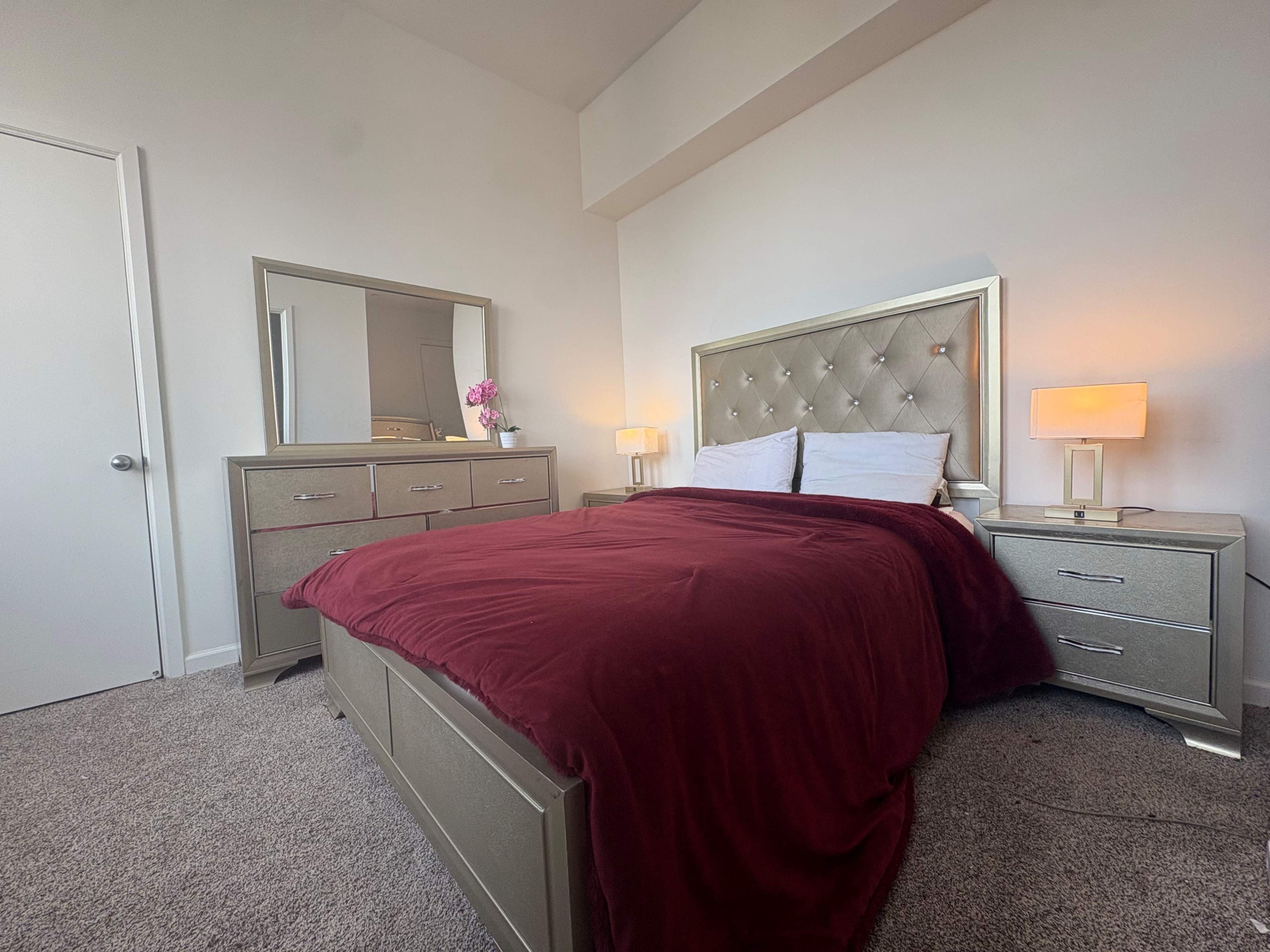 The image shows a well-furnished bedroom featuring a large bed with a burgundy blanket, a mirrored dresser, and a side table with a lamp.