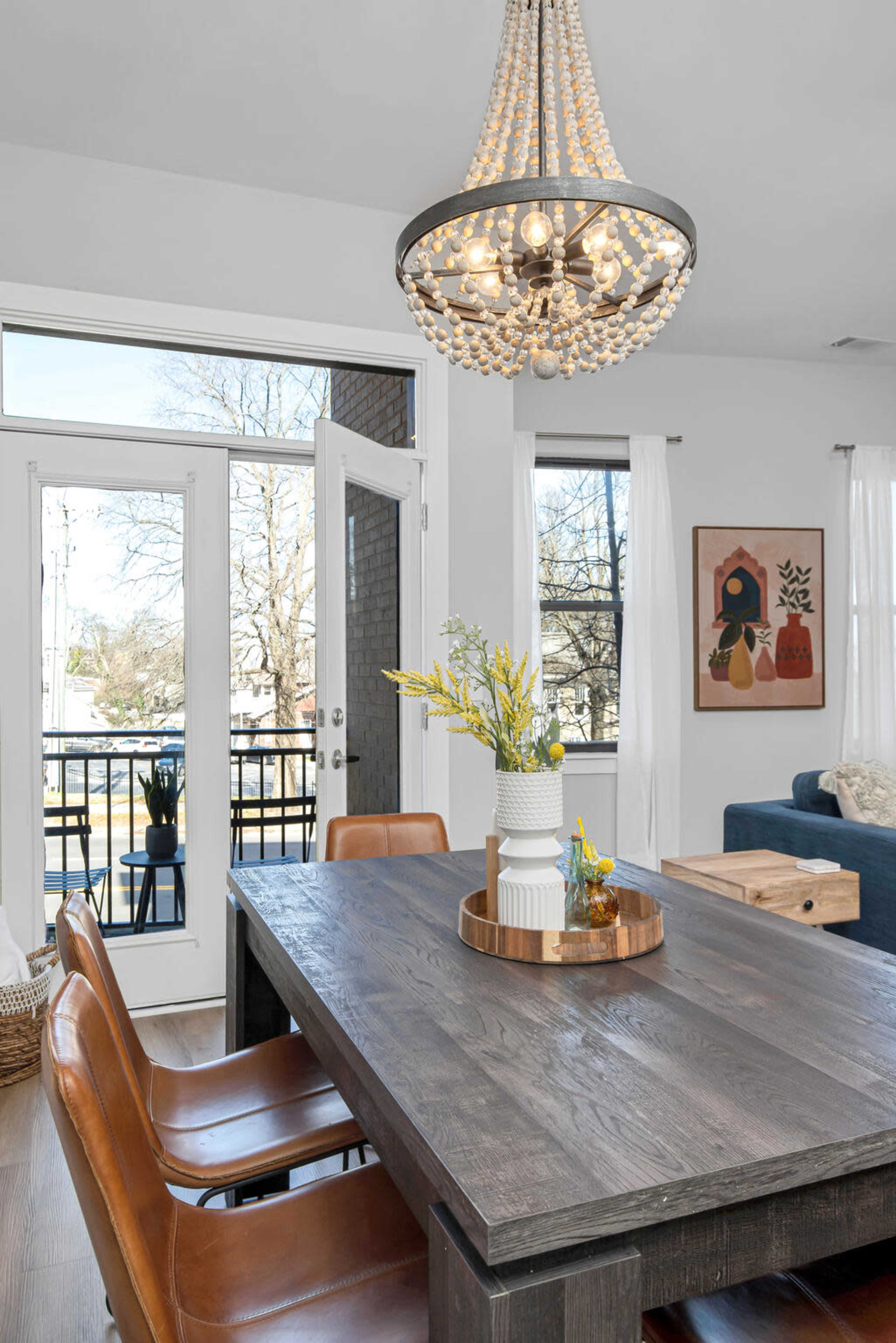 A modern dining area features a wooden table with leather chairs, a decorative centerpiece, and a chandelier, with large windows showcasing an outdoor view.
