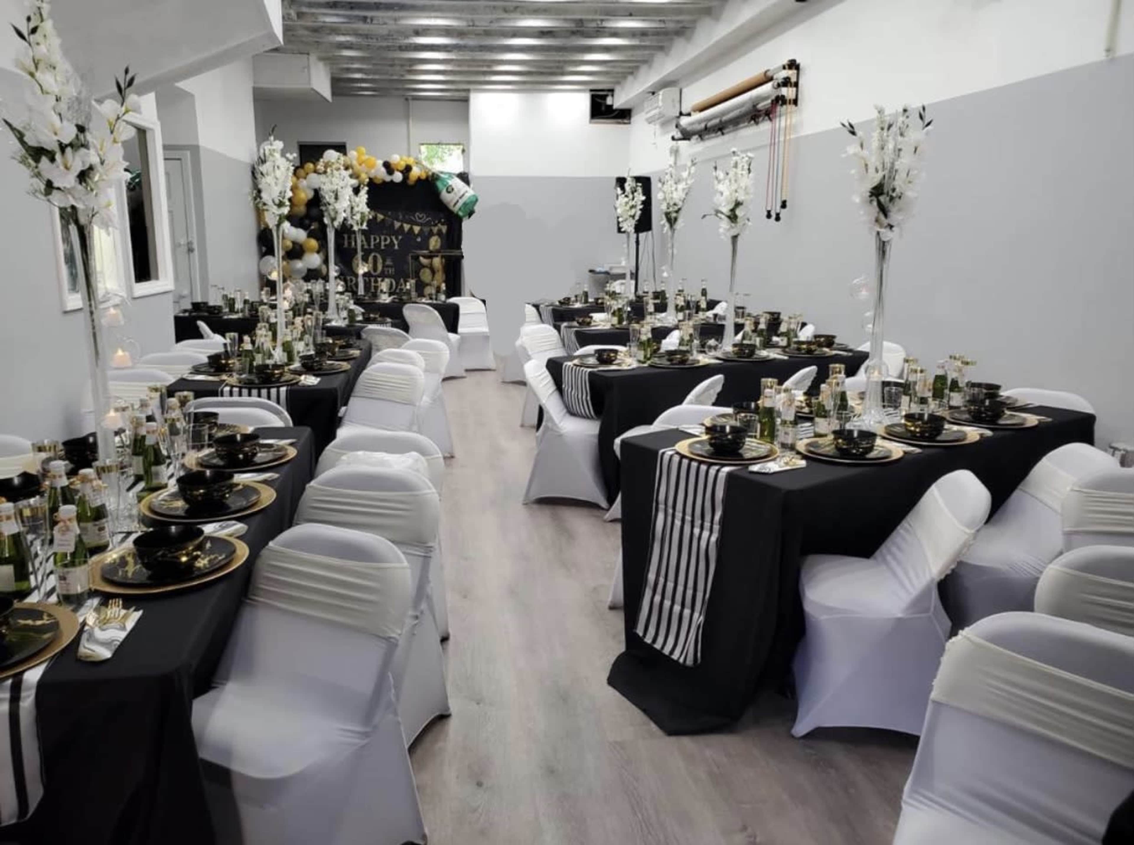 A banquet hall is set up with several elegantly arranged tables covered in black and white linens, featuring decorative centerpieces and tableware for a celebration.