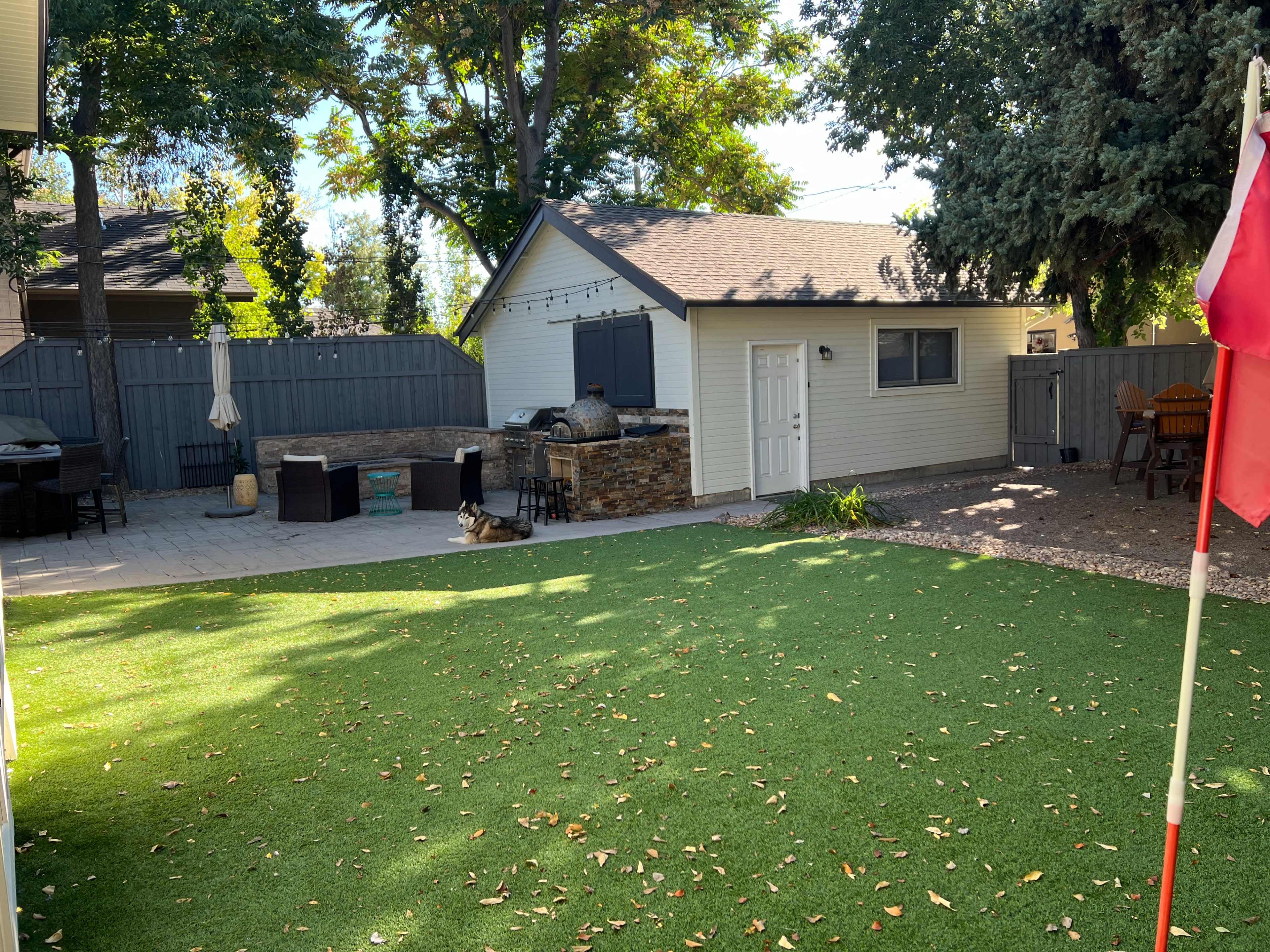 The image shows a landscaped backyard featuring a green artificial lawn, a wooden patio area, a small storage shed, and outdoor furniture.