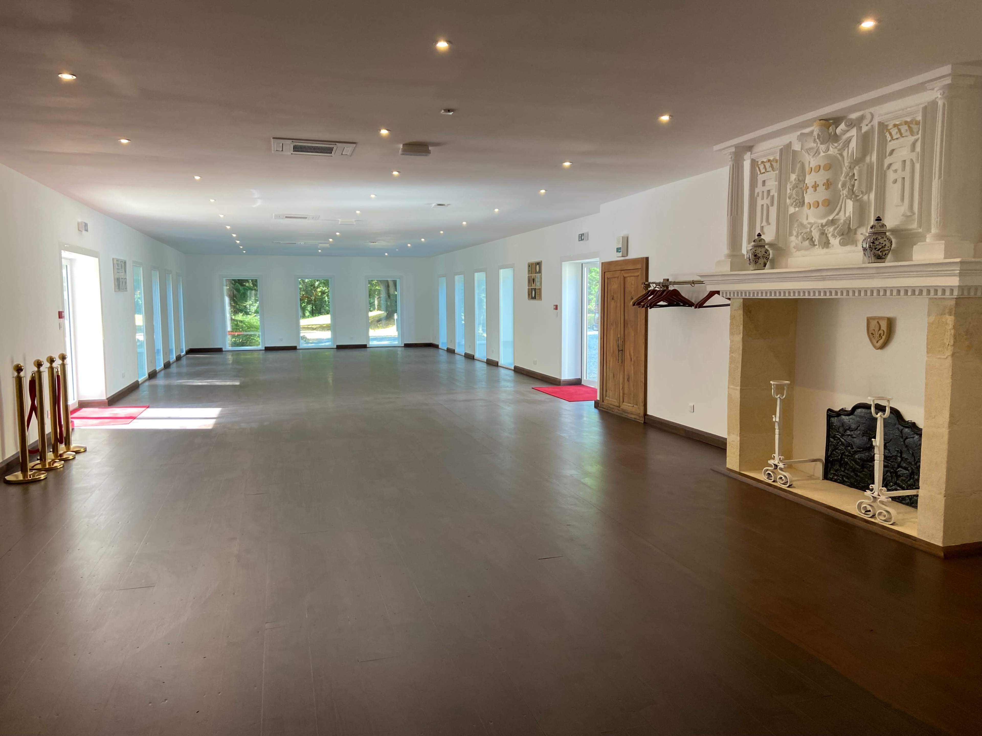 The image shows a spacious, well-lit hallway with polished wooden floors, featuring windows on one side and a decorative fireplace with a mantle on the opposite side.