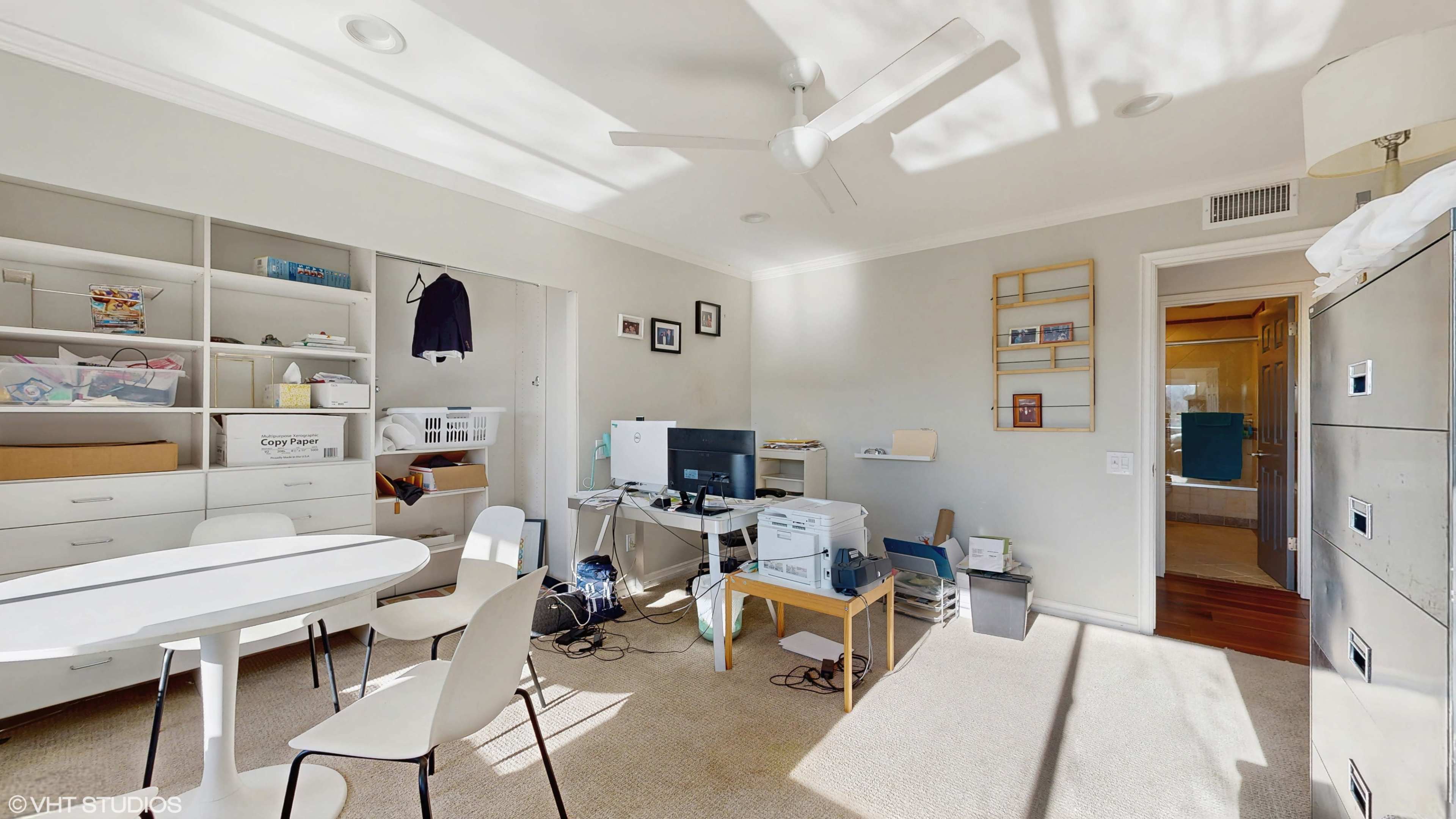 The image depicts a small office or workspace with a round table, several chairs, a computer setup, and various storage shelves containing organizational items.