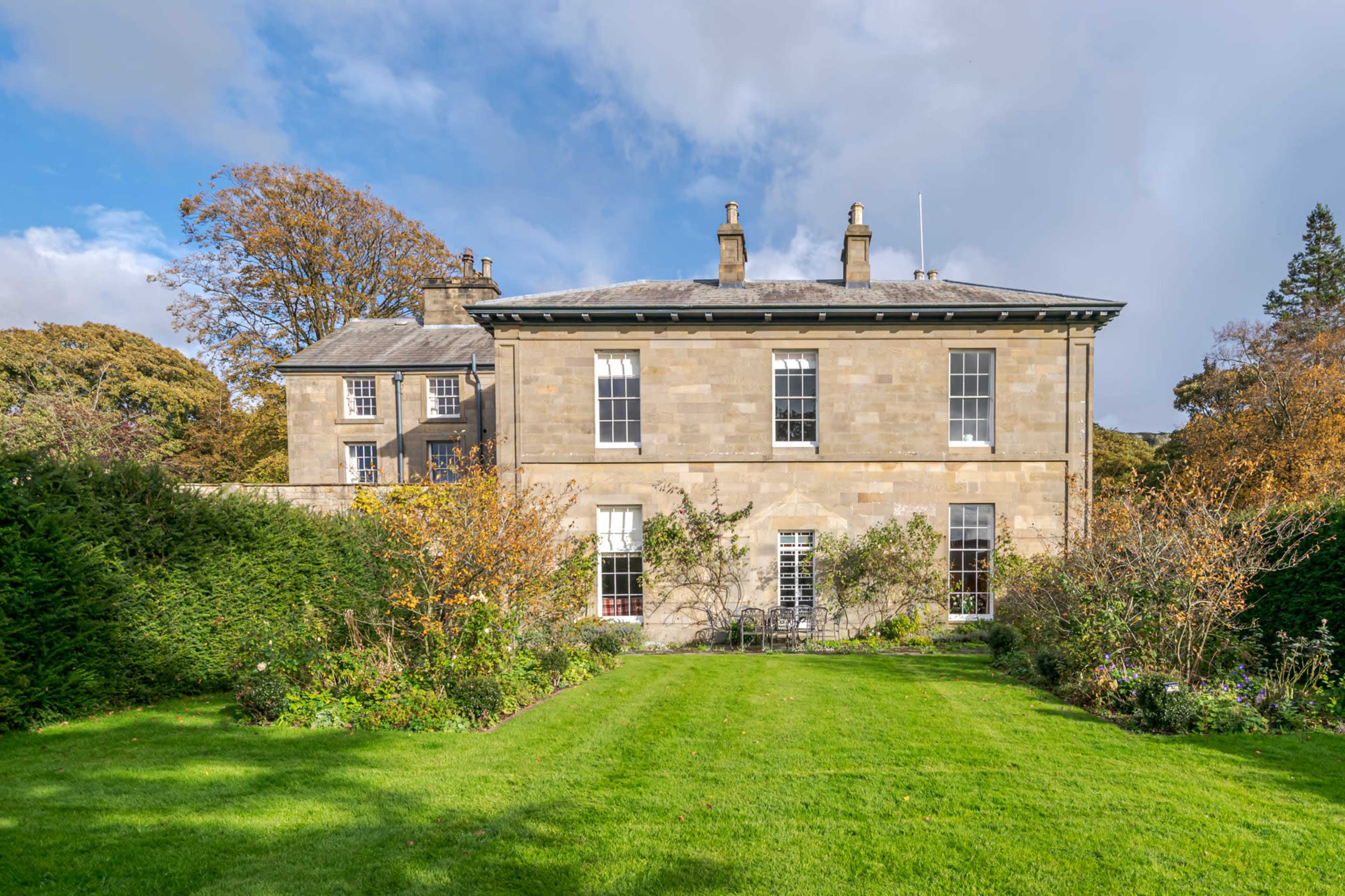 A two-story stone house with large windows surrounded by well-maintained gardens and trees.