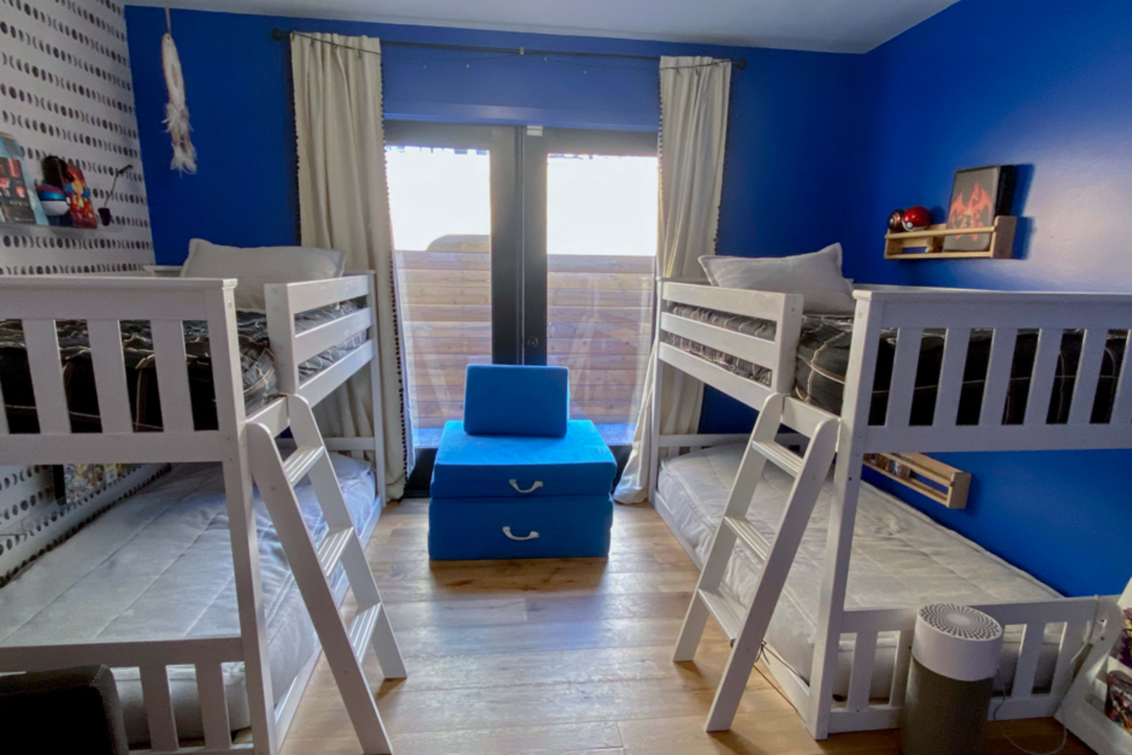The image depicts a children's bedroom featuring two sets of white bunk beds facing each other, with a blue storage cube in the center and a balcony visible through glass doors.