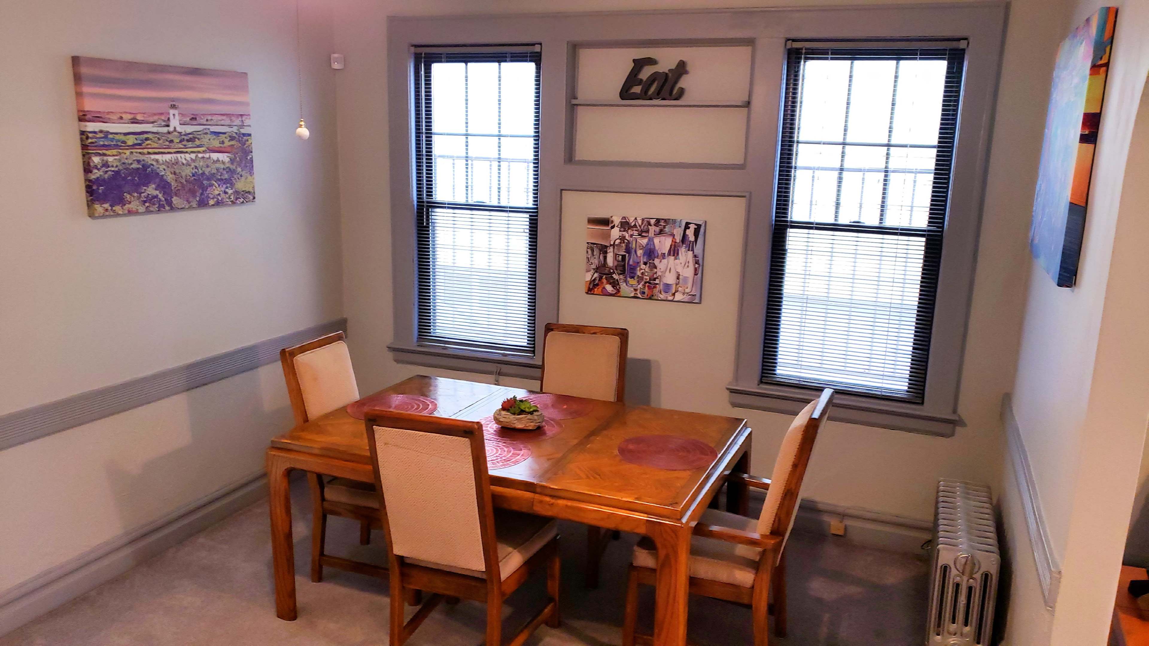 A wooden dining table with four chairs is set in a room featuring two windows, a wall-mounted sign that says "Eat," and various framed photos and artwork.
