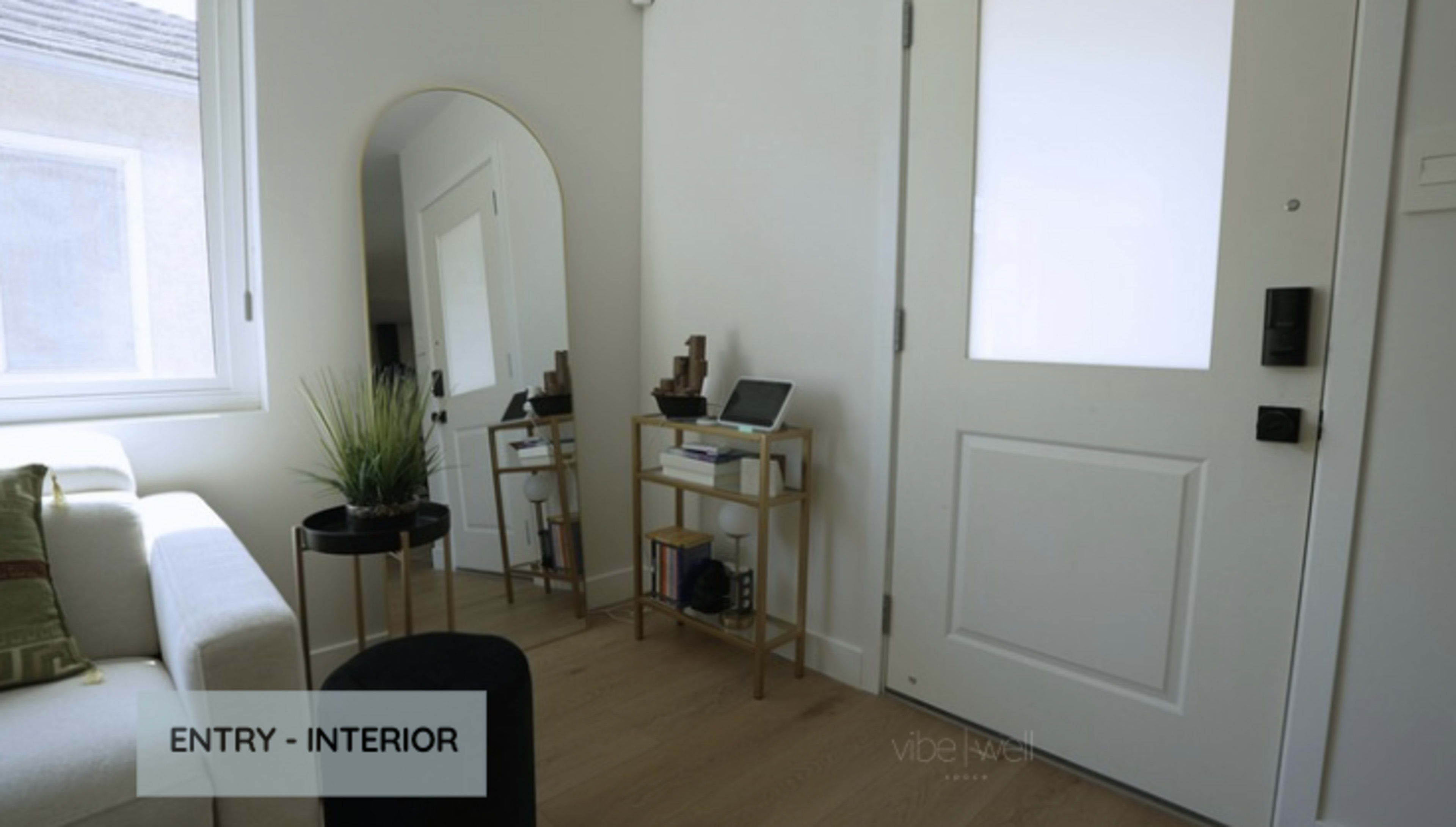The image shows an entryway with a door on the right, a round mirror standing on the left, and a small console table with decorative items and a tablet.