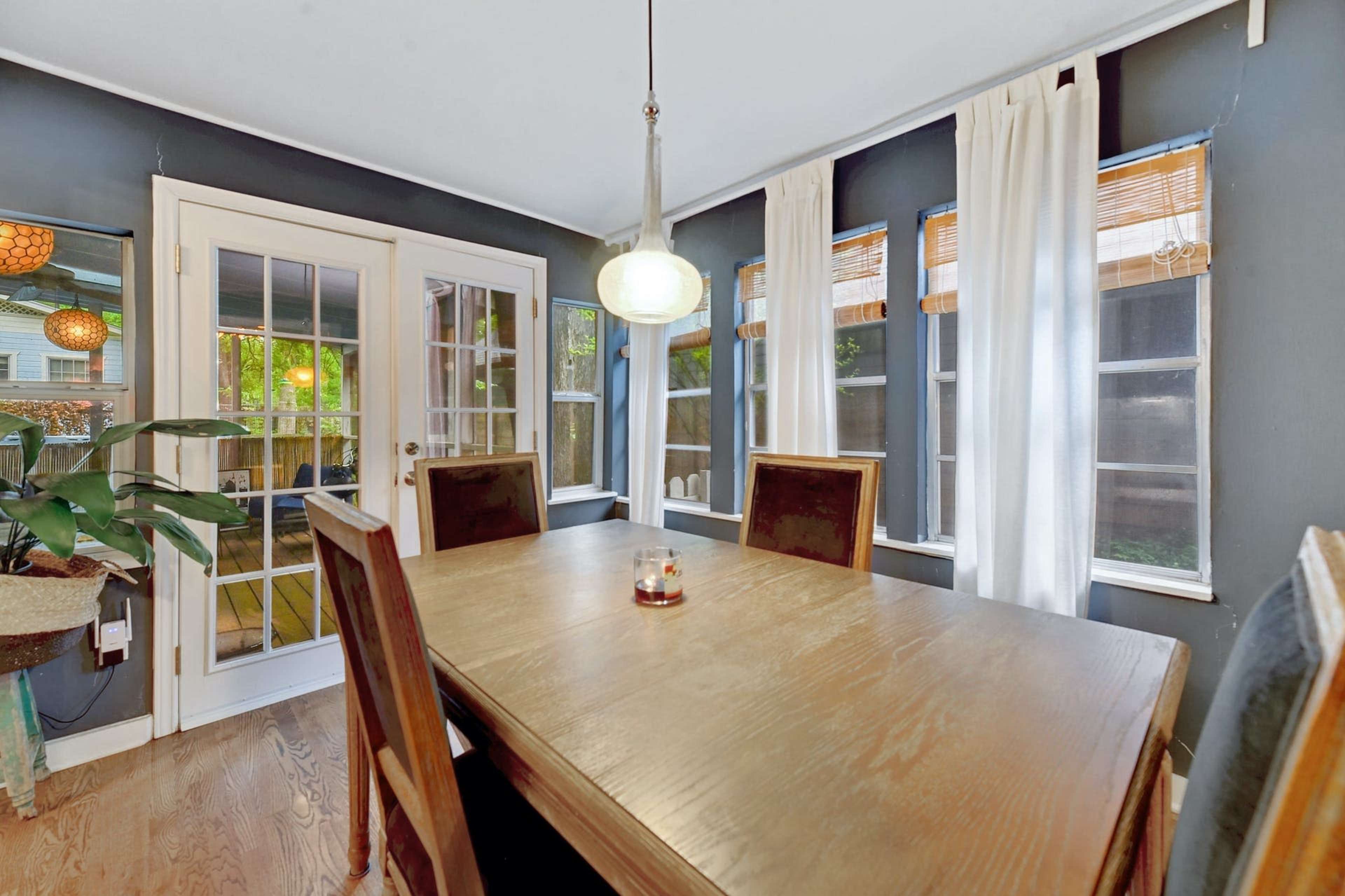 A dining table with wooden chairs is situated near large windows adorned with light curtains in a room with dark walls.