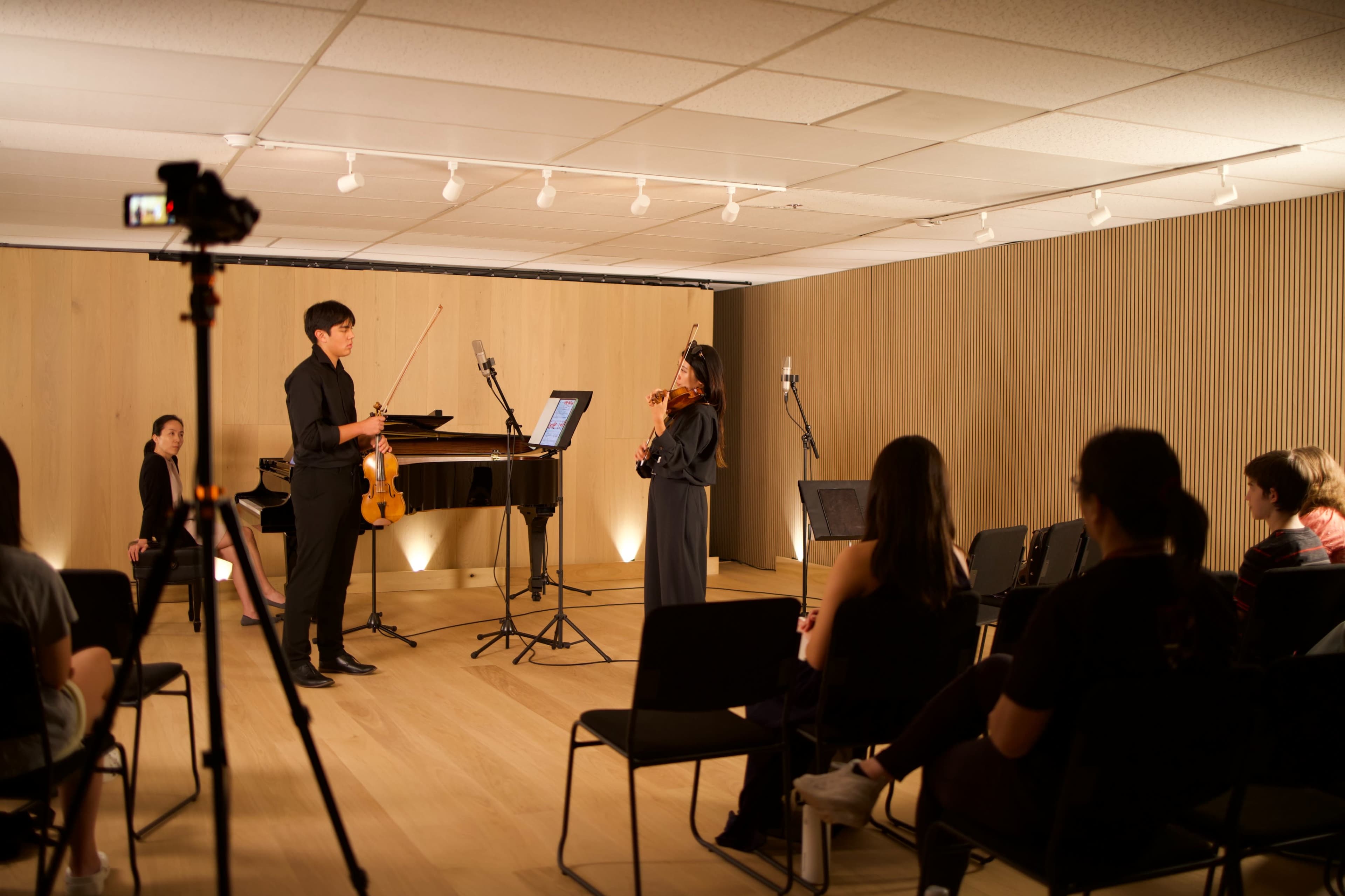 A violinist stands in front of a piano while a small audience watches in a well-lit performance space.