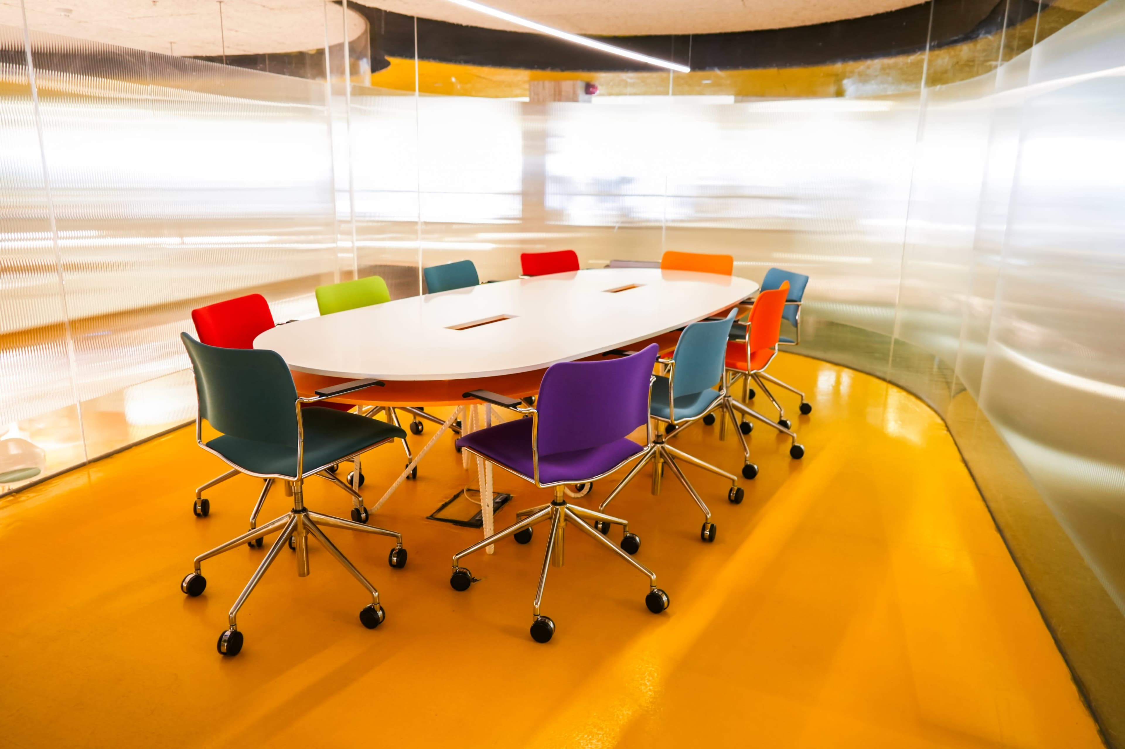 A round conference table with colorful chairs is located in a glass-walled meeting room with an orange floor.