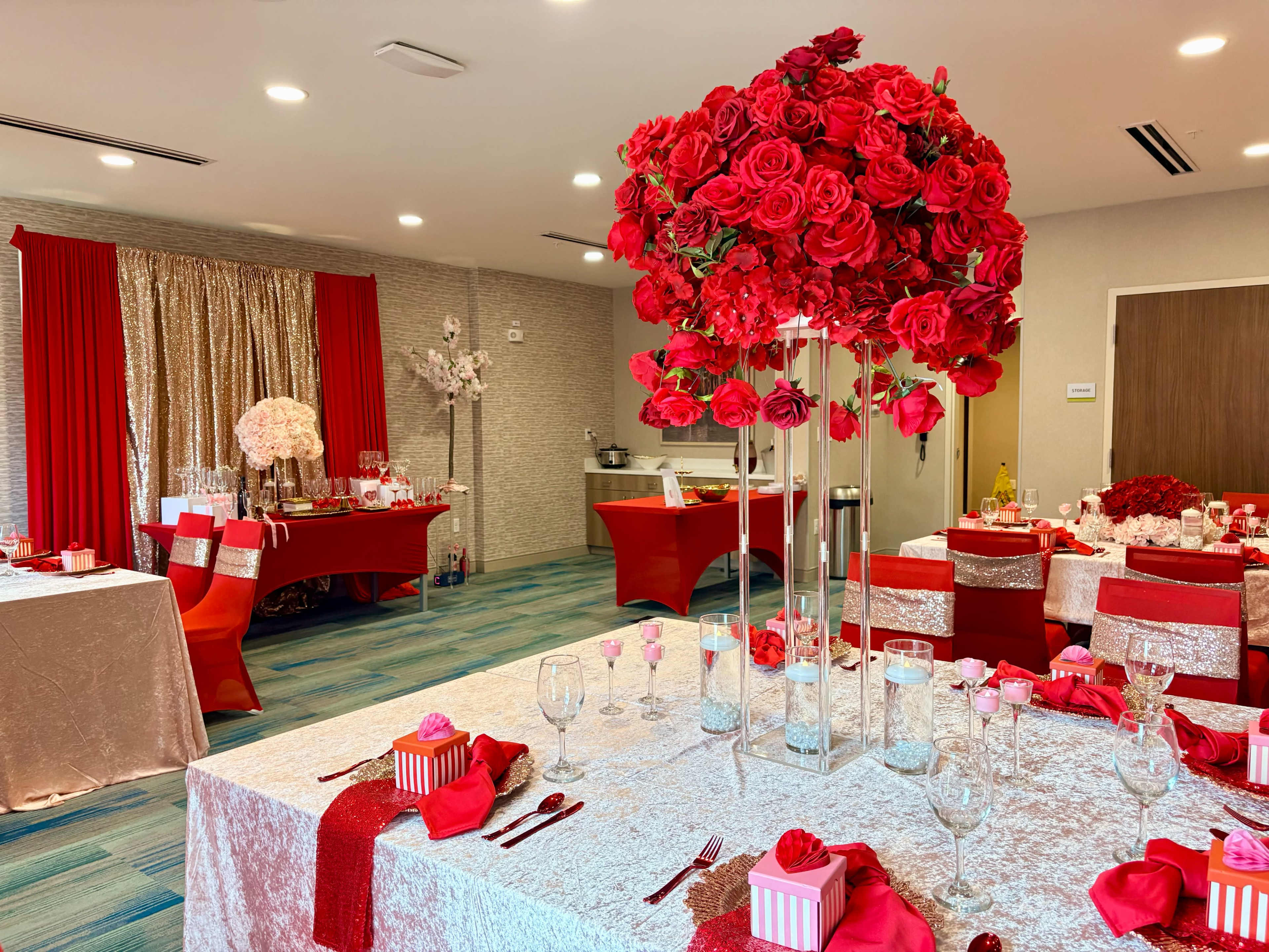 The image shows an elegantly decorated banquet room with tables adorned in red and silver, featuring tall floral arrangements of red roses.