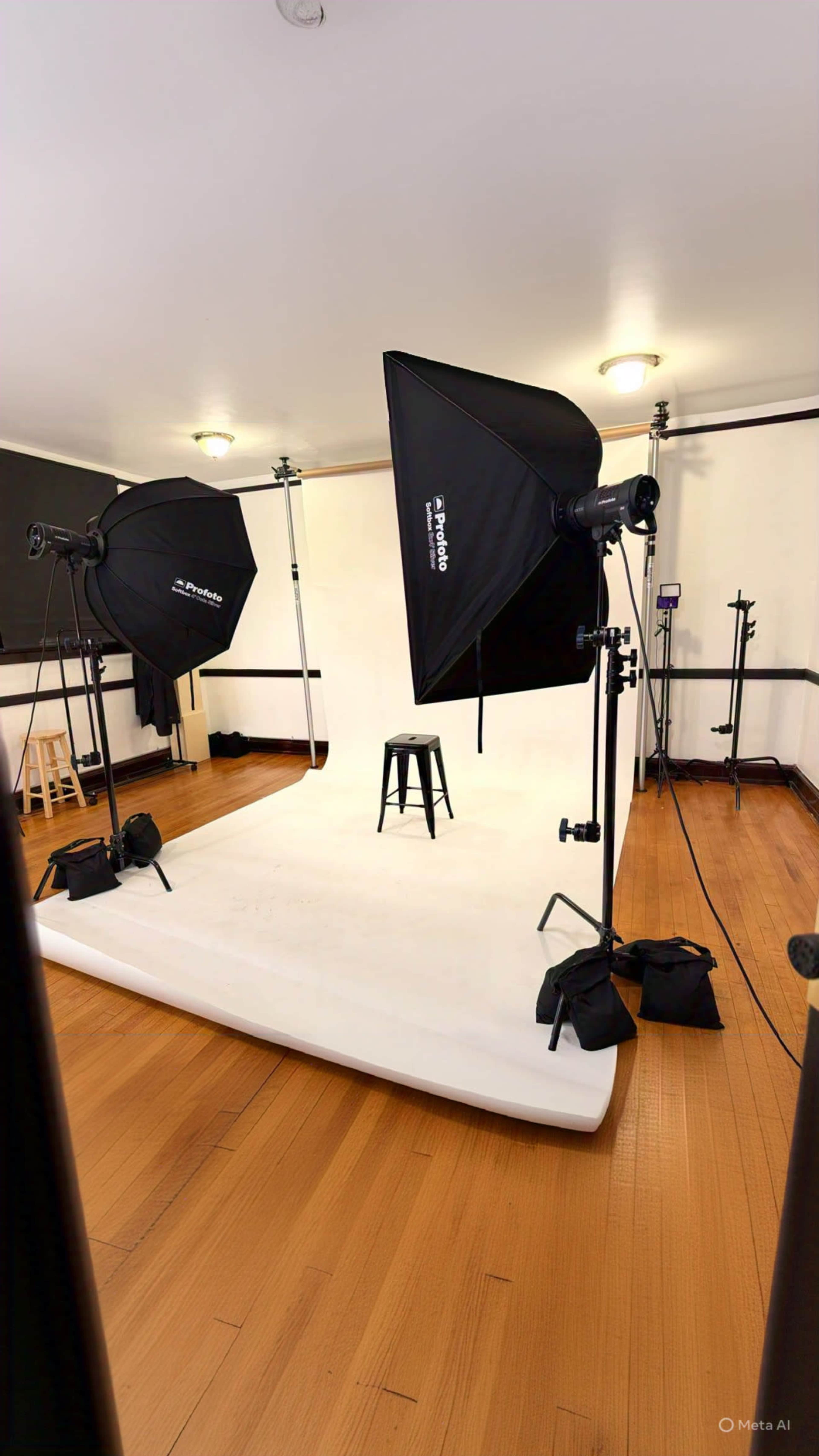 The image shows a photo studio setup with two large softbox lights illuminating a white backdrop and a black stool in the center.