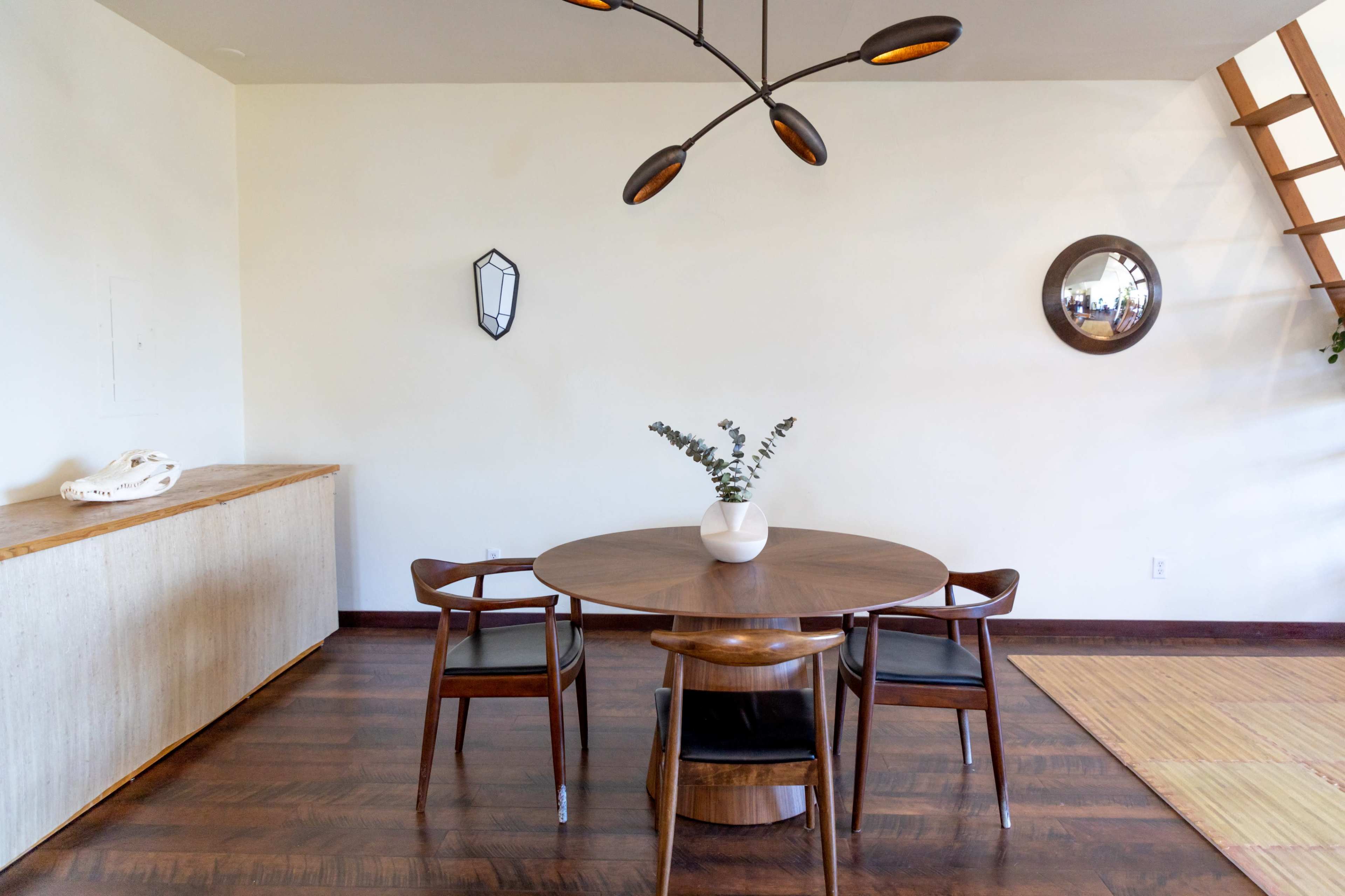 The image shows a minimalist dining area featuring a round wooden table surrounded by three chairs, a decorative plant in a vase, and mid-century modern lighting fixtures.