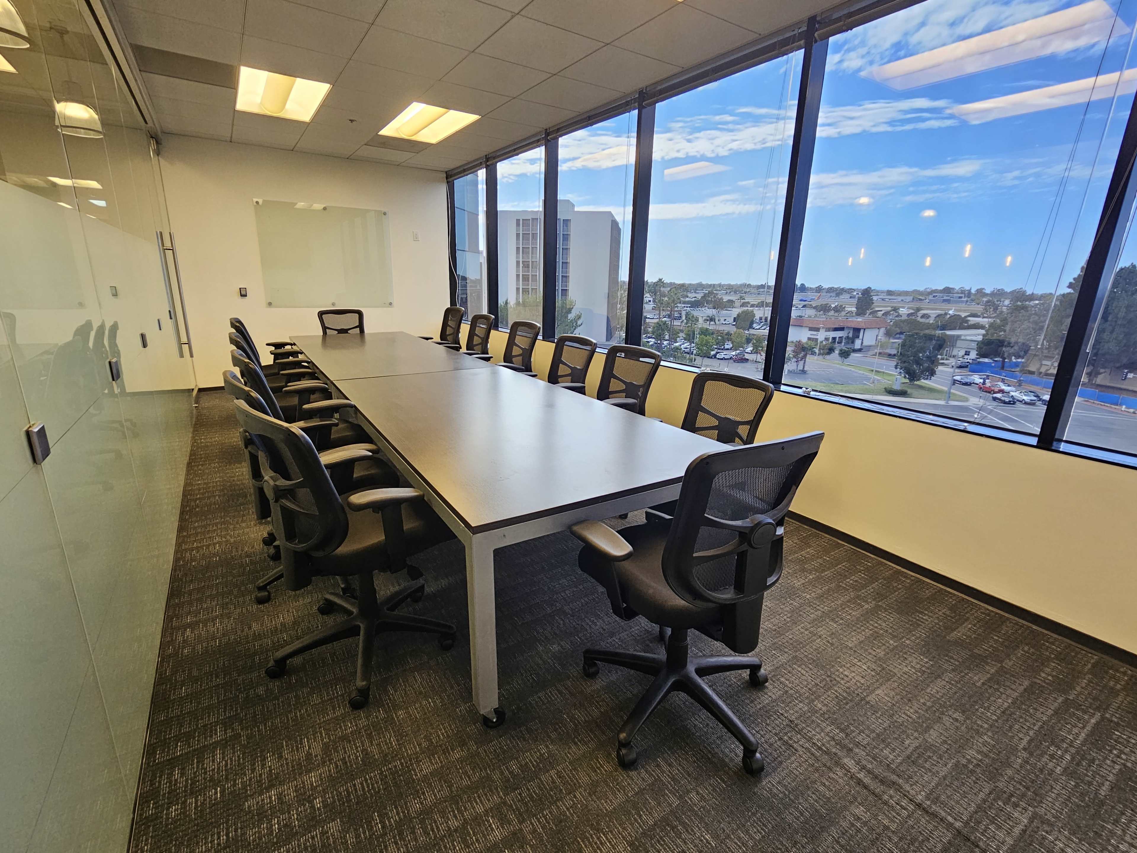 A large conference room features a long table surrounded by ergonomic black chairs, with windows offering a view of the skyline and landscape outside.