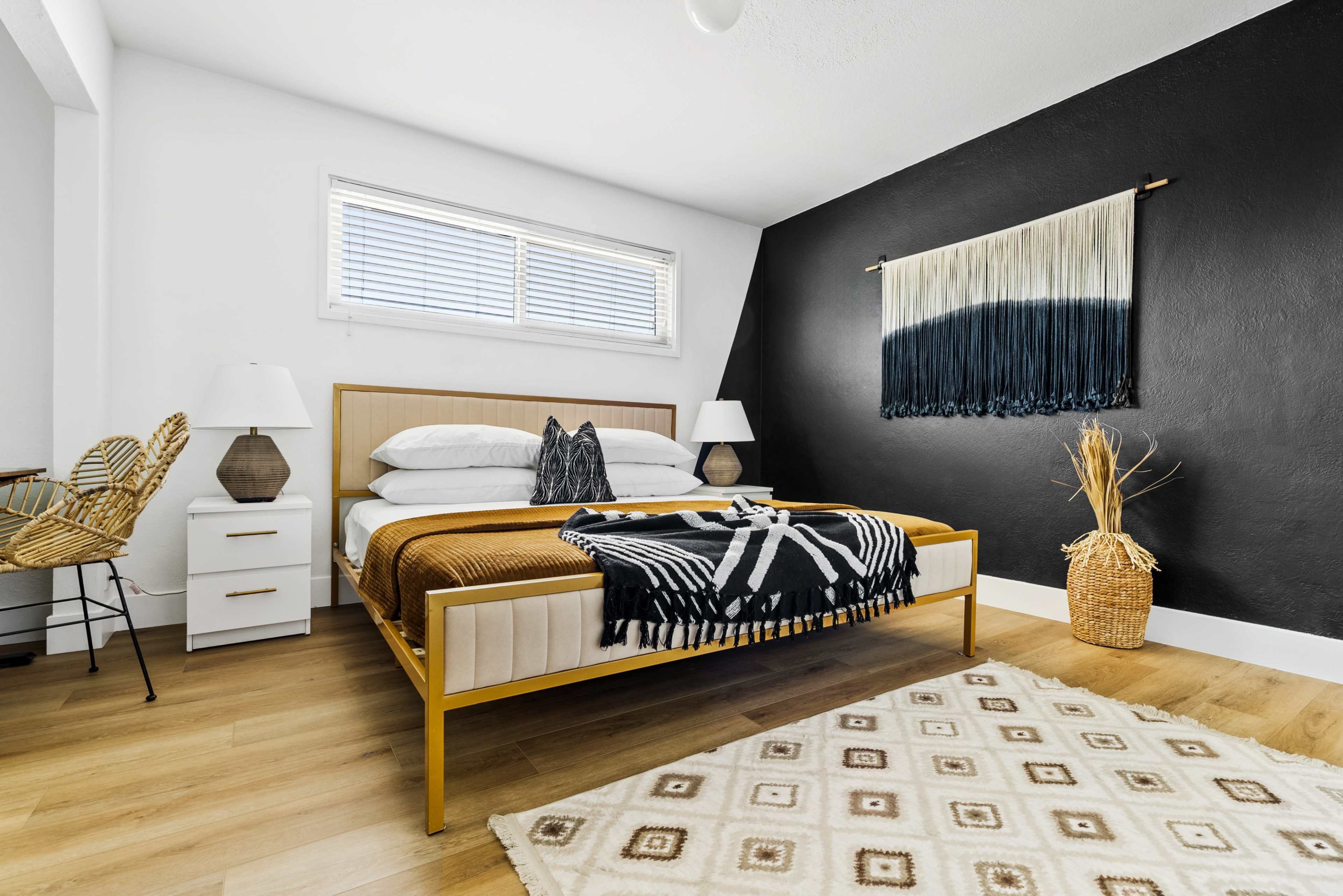 The image shows a modern bedroom featuring a wooden bed with a patterned blanket, two bedside lamps, a woven chair, and a wall adorned with a textile hanging.