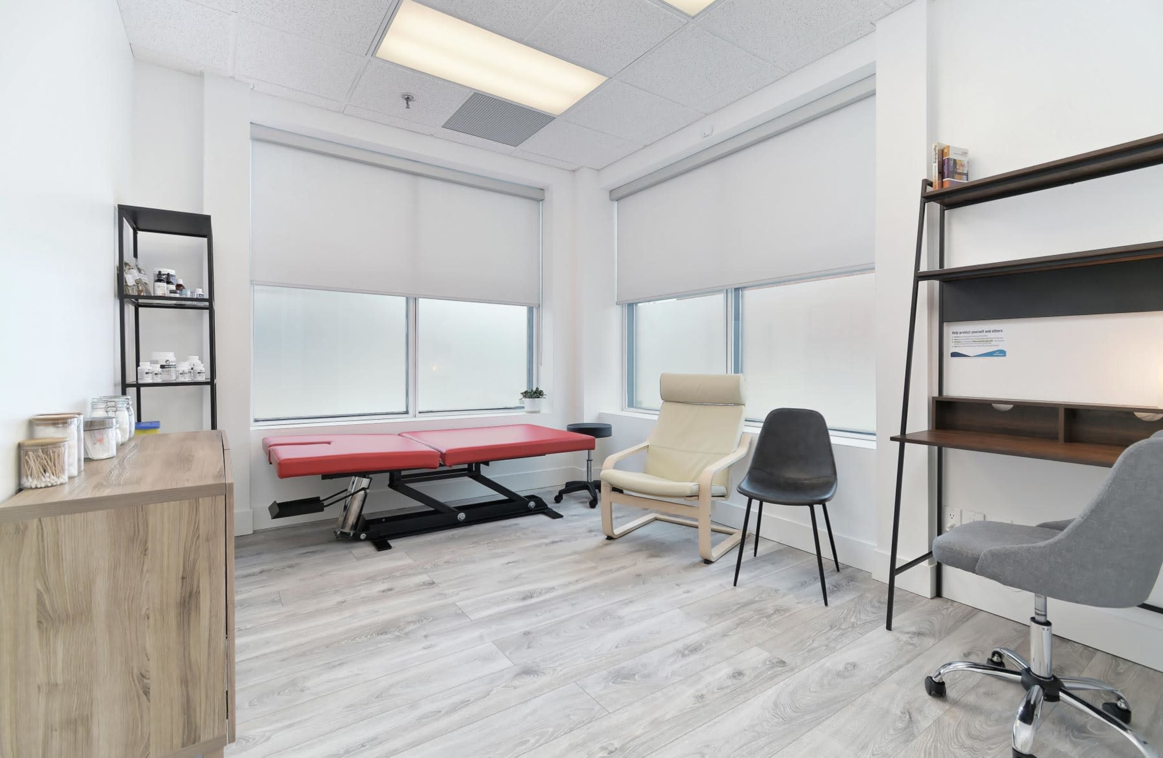 A minimally furnished therapy room with a massage table, a chair, and a small shelf, featuring large windows that allow natural light.