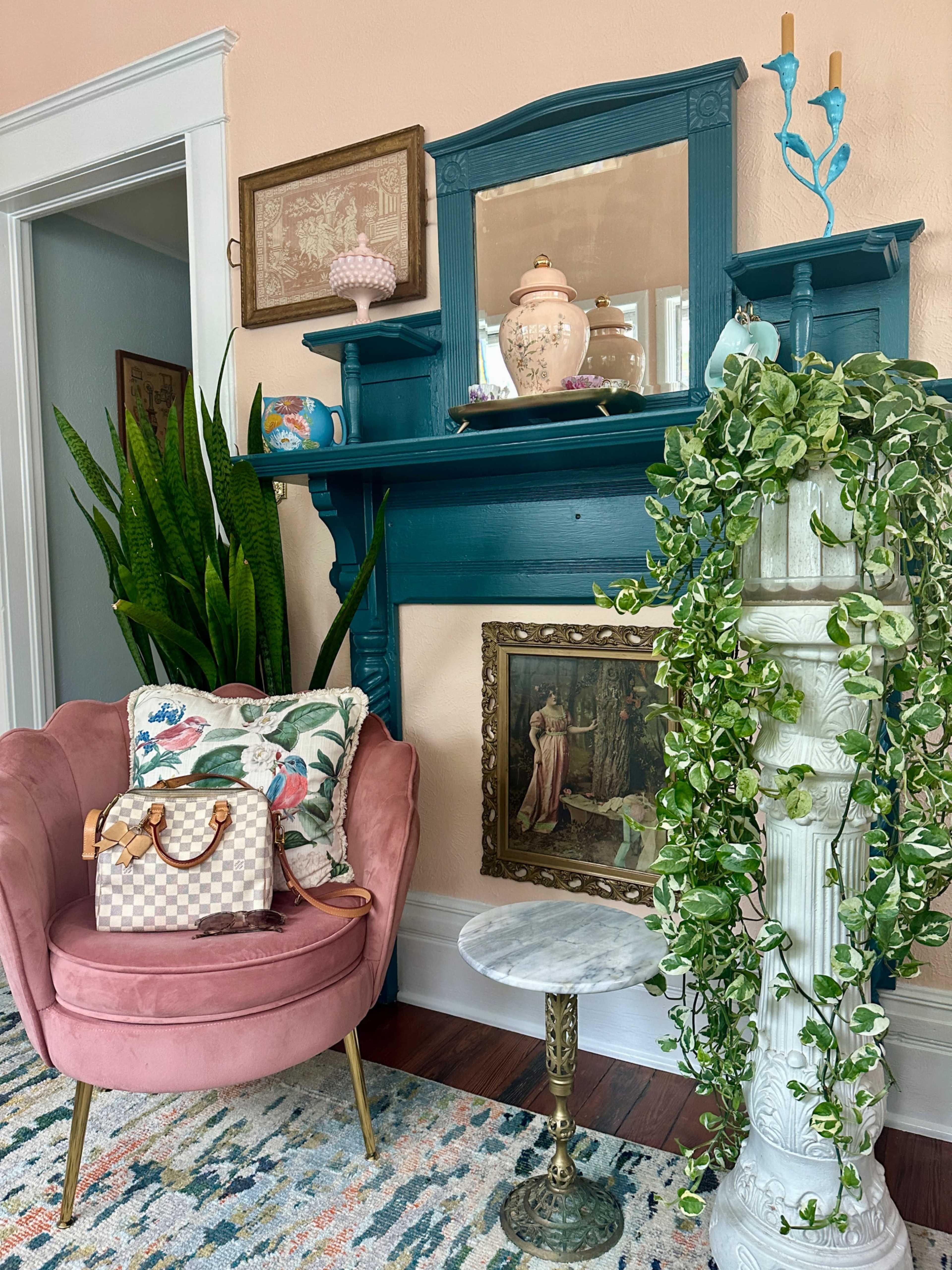 A pink armchair with floral pillows sits next to a tall green plant and a decorative white pedestal, all beside a blue mantel adorned with framed art and accessories.