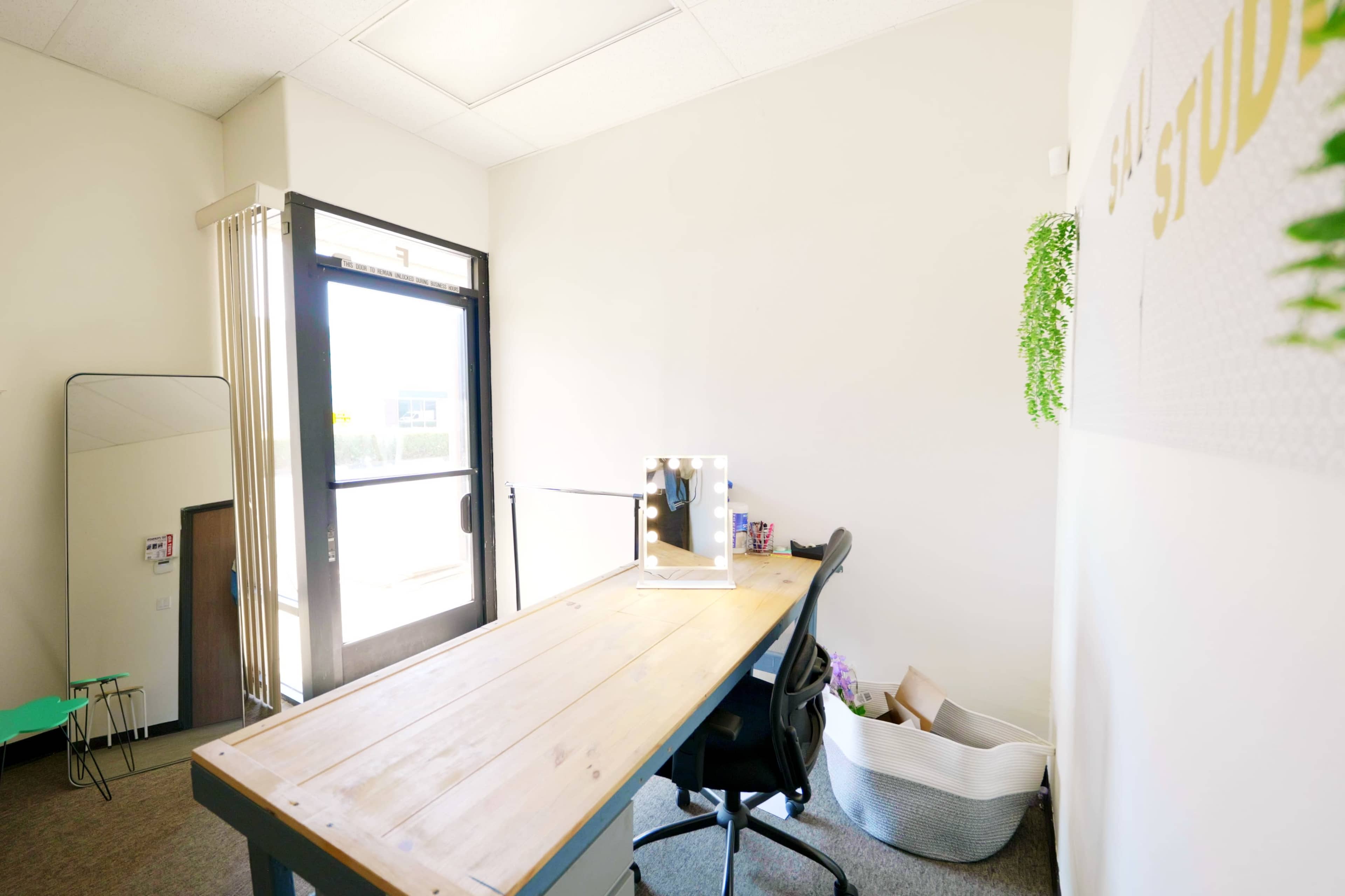 A desk sits in a bright, minimalist office space with large windows, and a potted plant is visible on the wall.