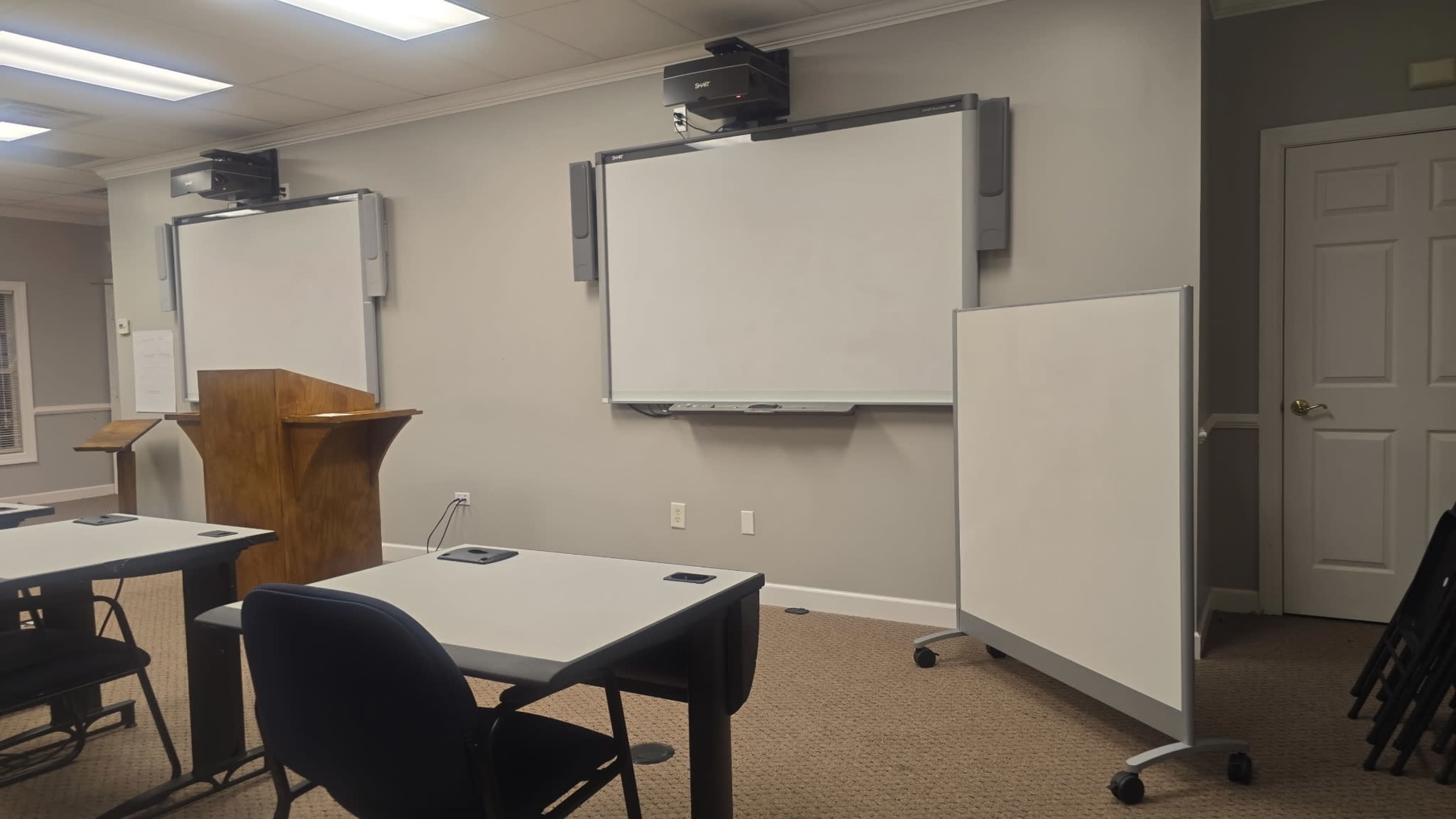 The image shows a classroom setting with two whiteboards, a wooden podium, and several tables and chairs arranged for instructional activities.