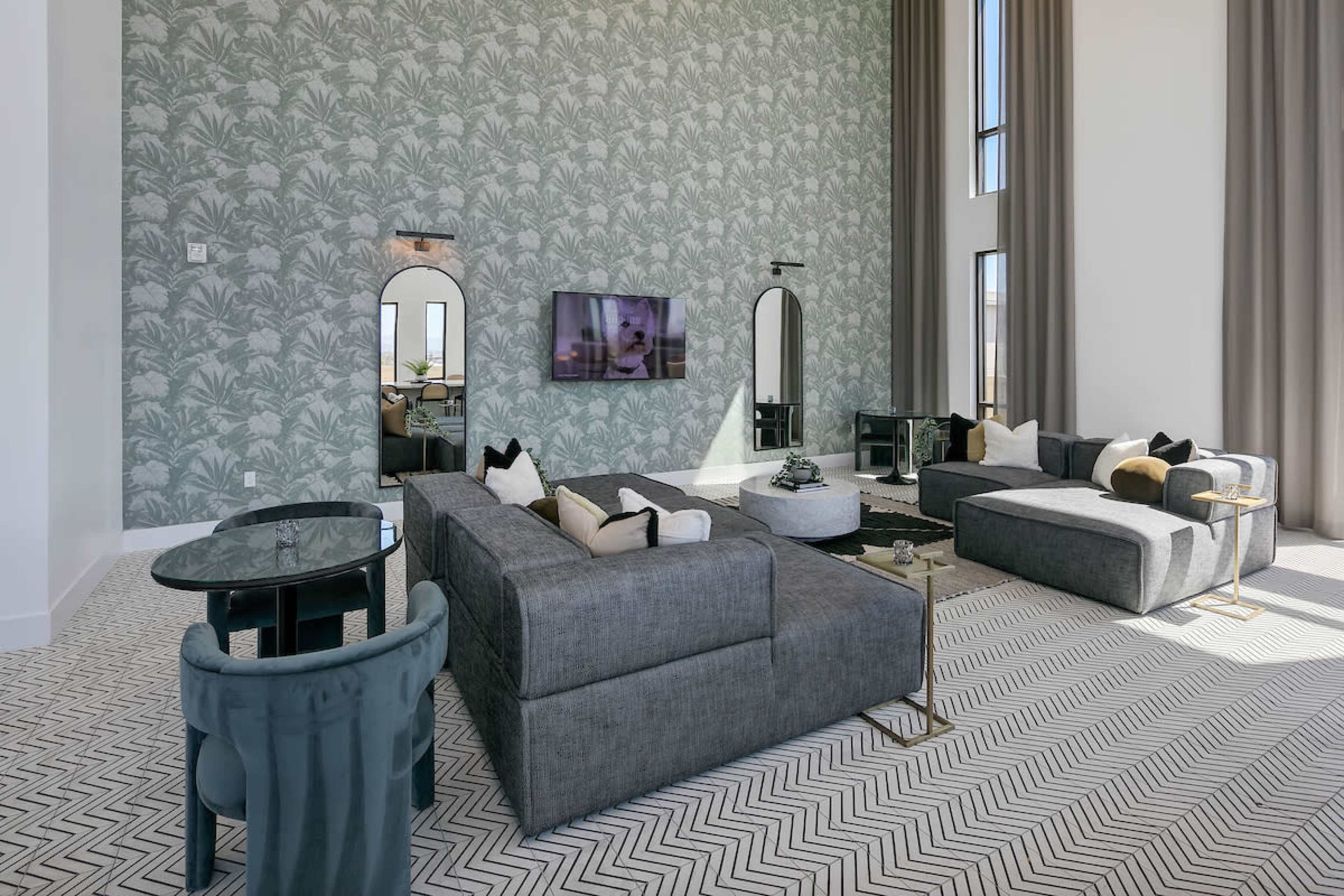 The living room features a light gray sectional sofa, a round black table, and large windows, all set against a patterned green wallpaper.