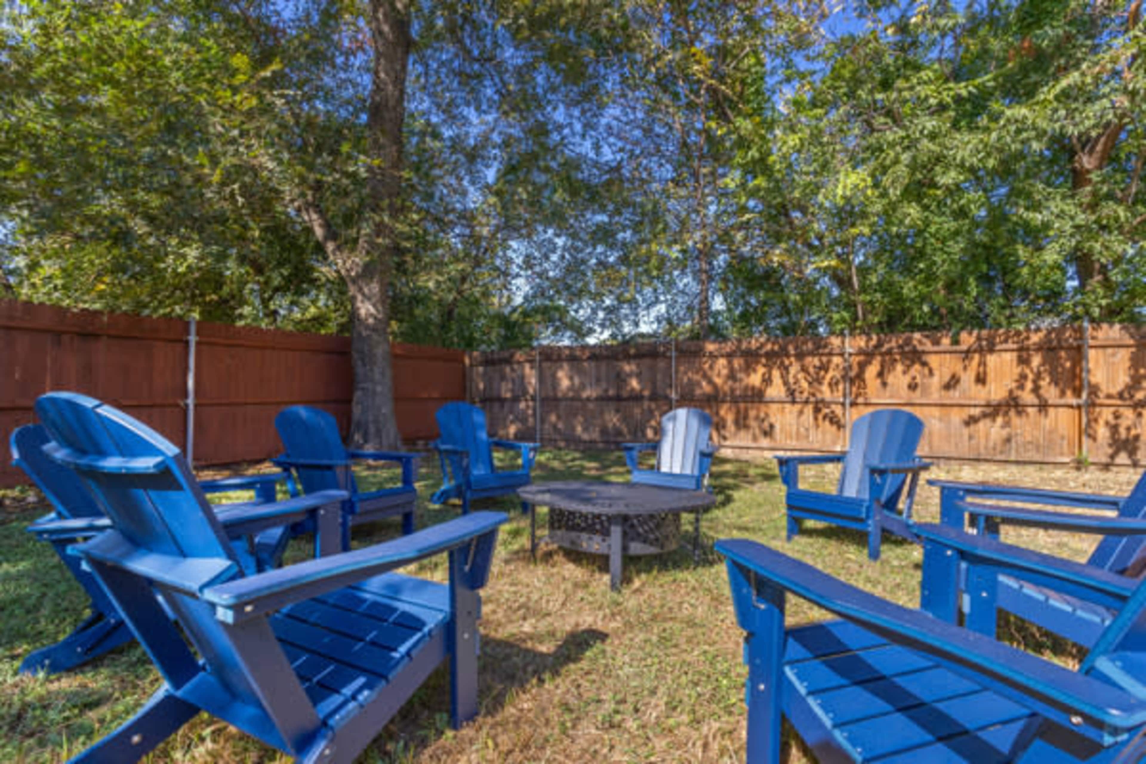 The image shows a backyard setting with several blue Adirondack chairs arranged in a circle around a fire pit, surrounded by trees and a wooden fence.