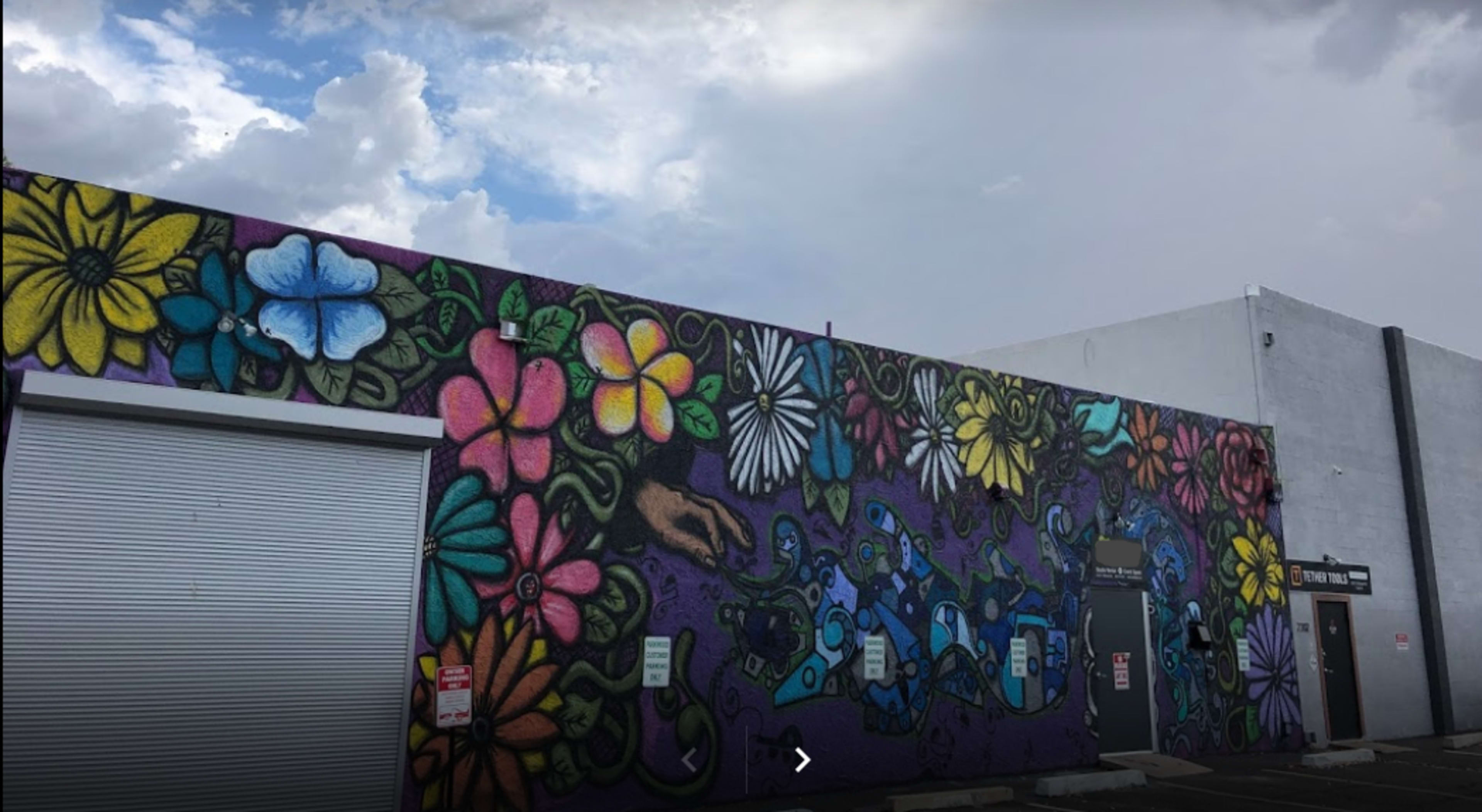 A colorful mural featuring various flowers and a hand is painted on the side of a building, under a partly cloudy sky.