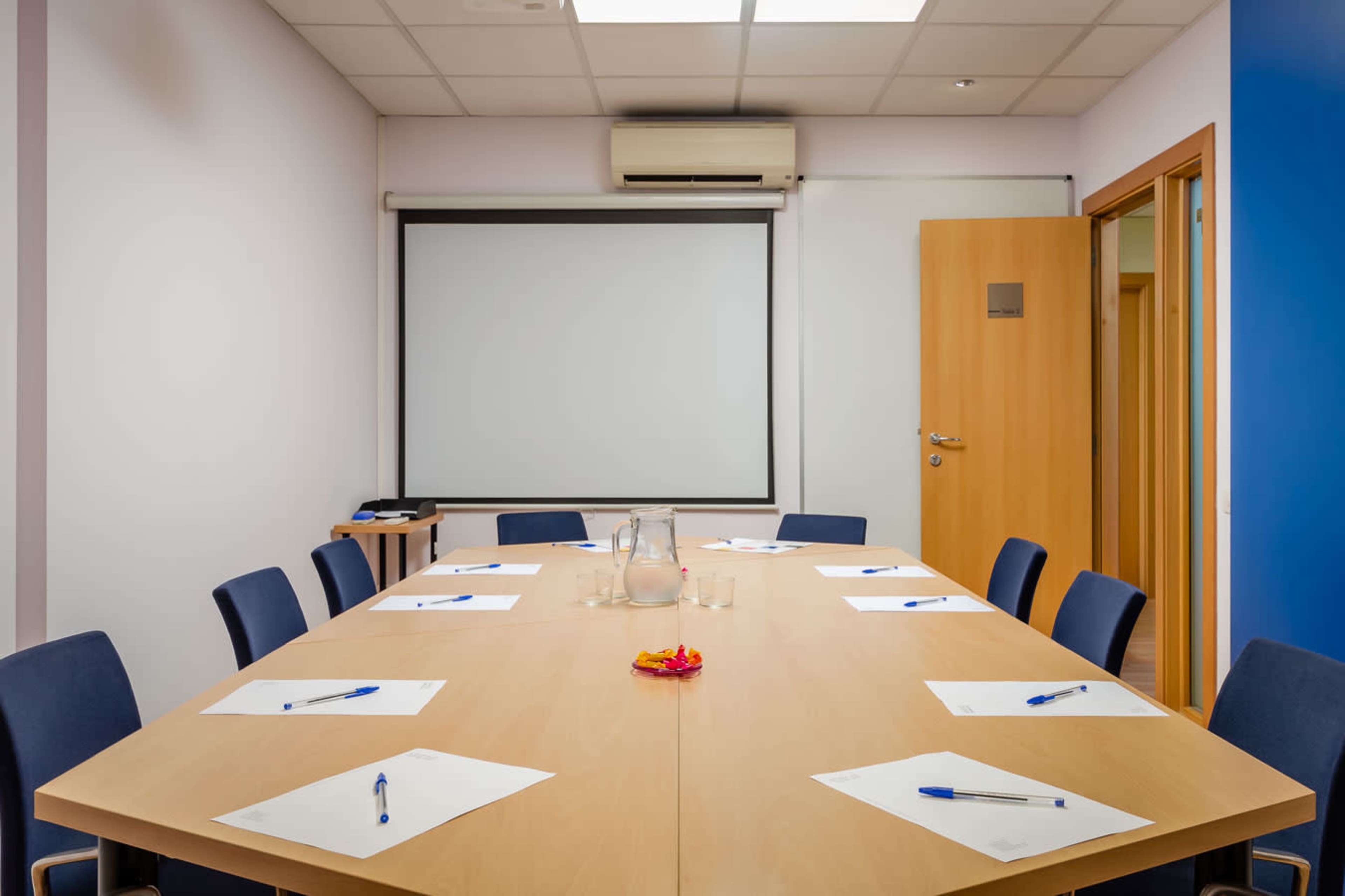 A conference room features a large table surrounded by chairs, with a projector screen on one wall and stationery laid out for meetings.