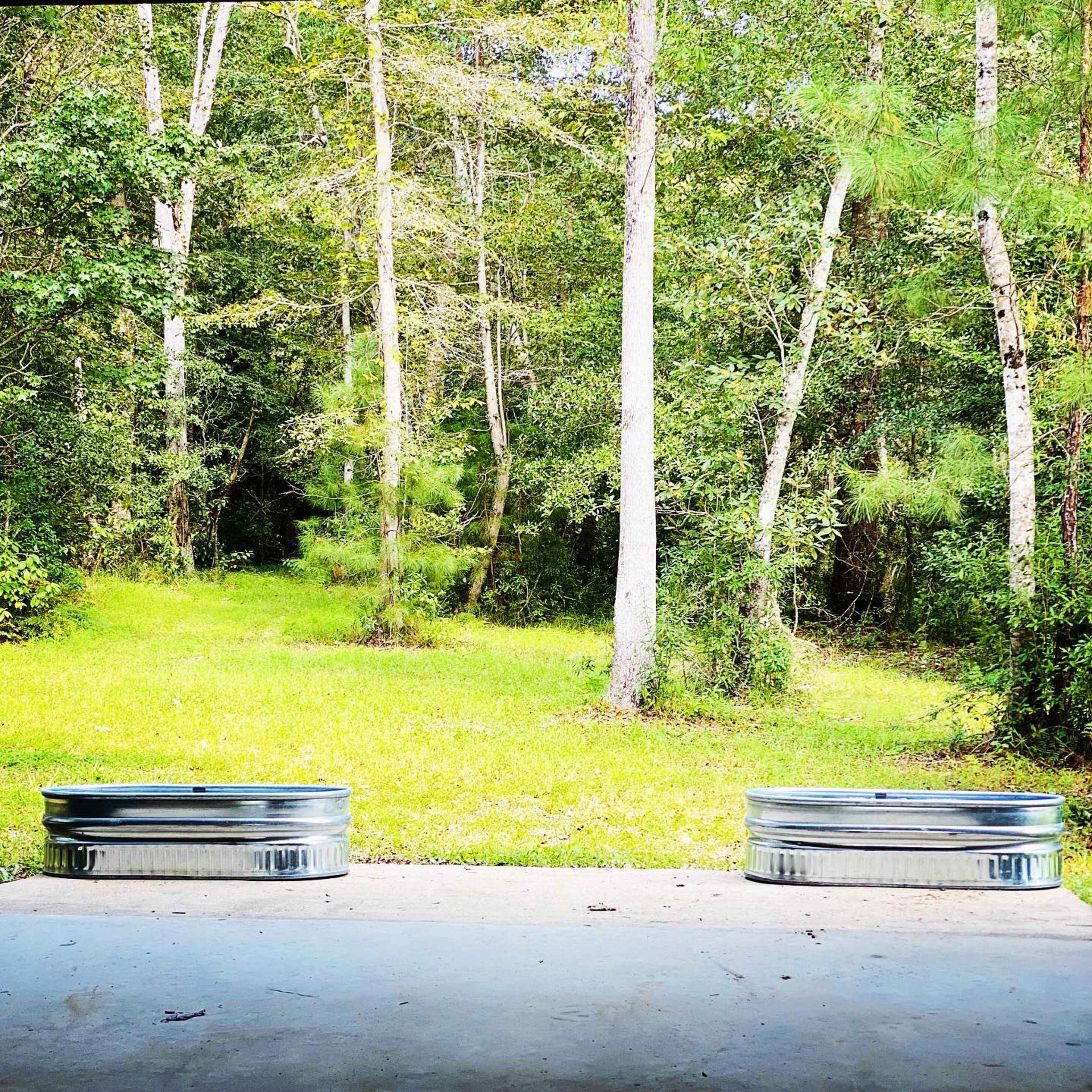 Two metal planters placed on a concrete surface facing a lush, green forest with tall trees.