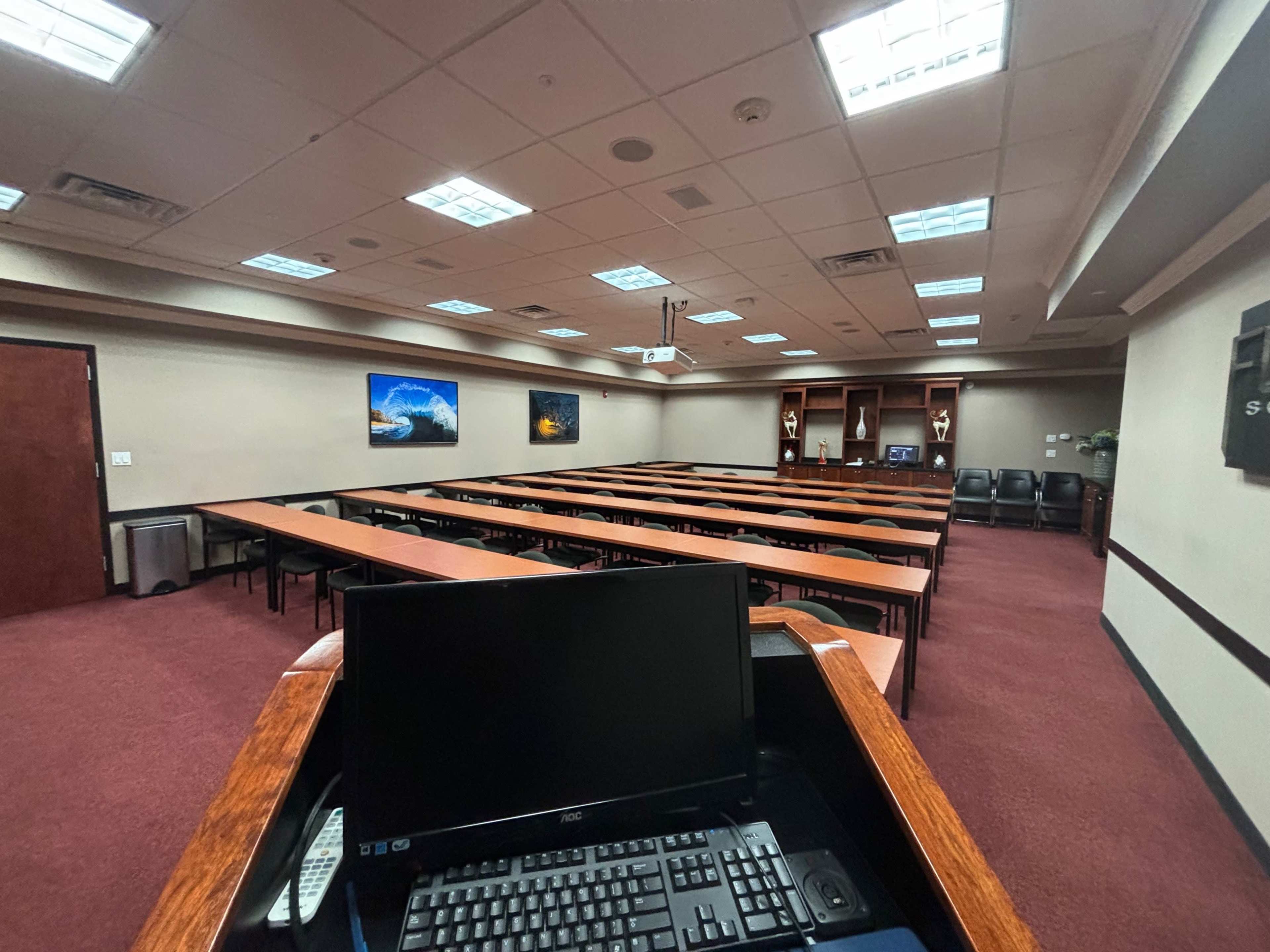 A training room with rows of wooden tables and chairs, featuring a computer at the front and artwork on the walls.