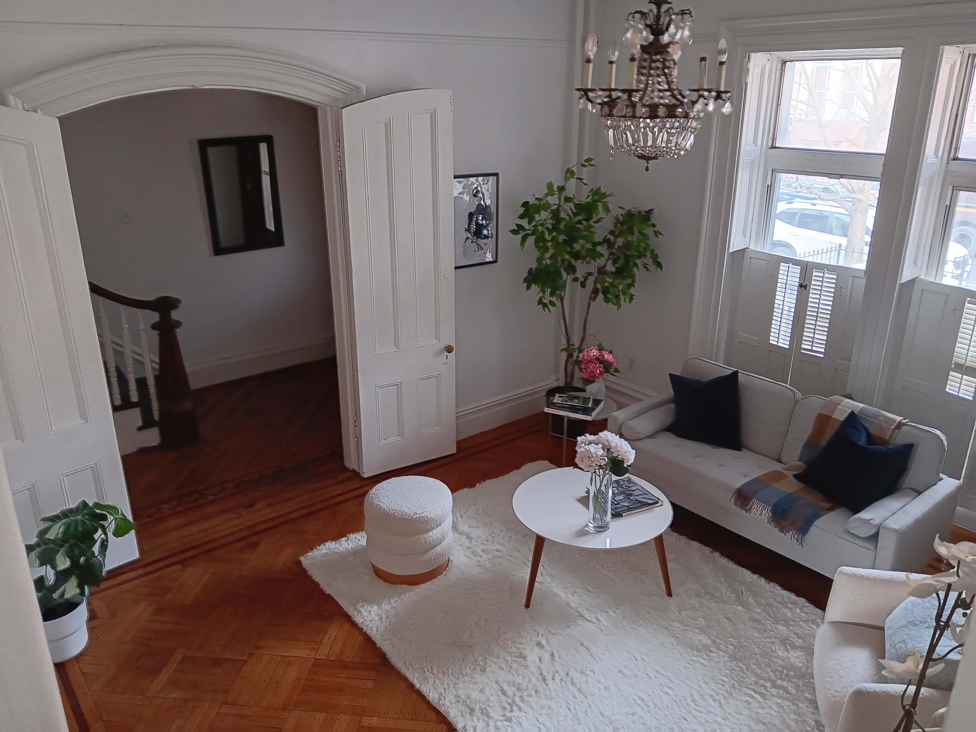 A well-lit living room features a white sofa, a round coffee table, and a plush area rug, with decorative plants and artwork enhancing the space.