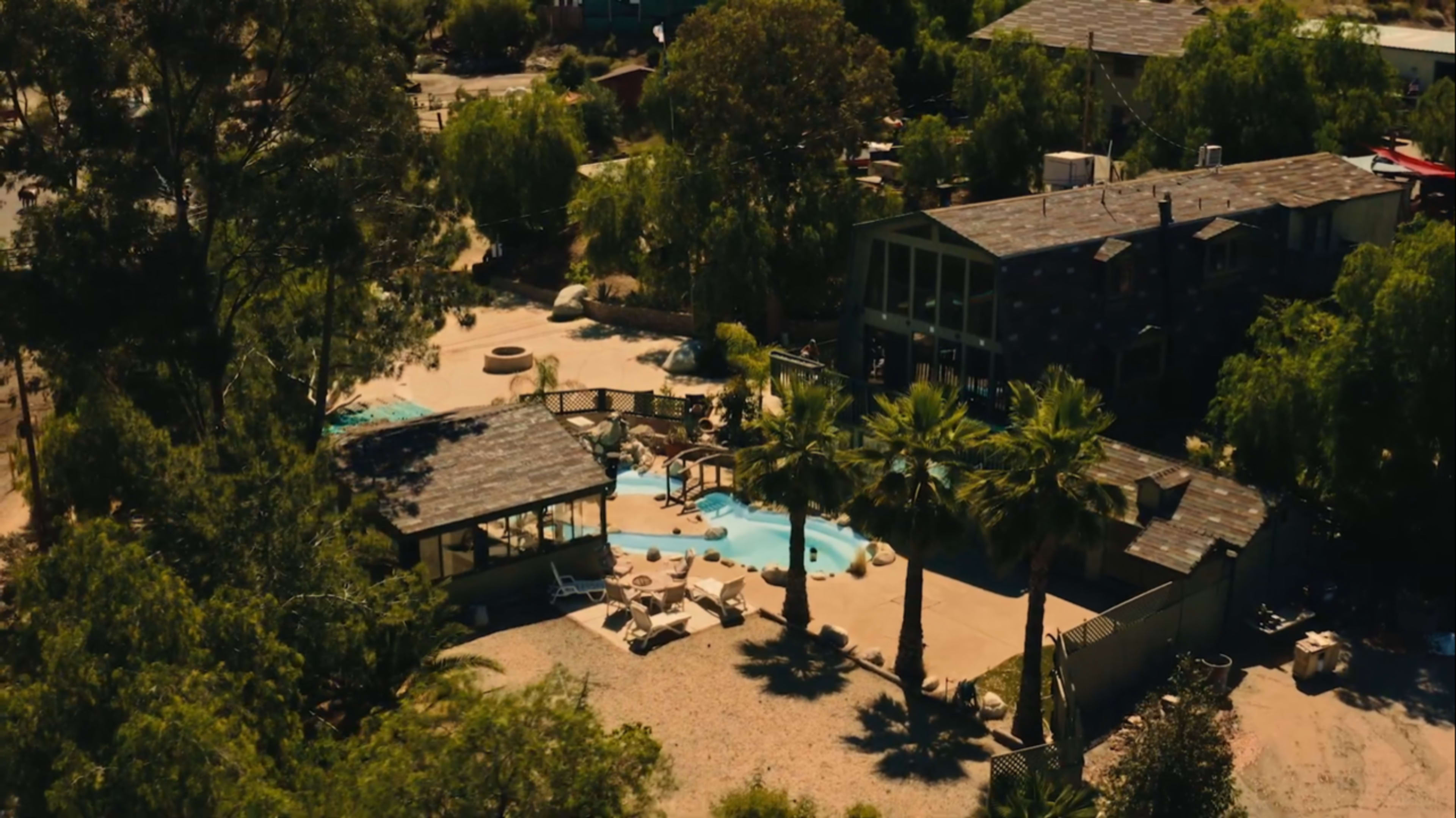 An aerial view of a spacious outdoor area with a swimming pool surrounded by palm trees, lounge chairs, and several buildings in a greenery-filled setting.