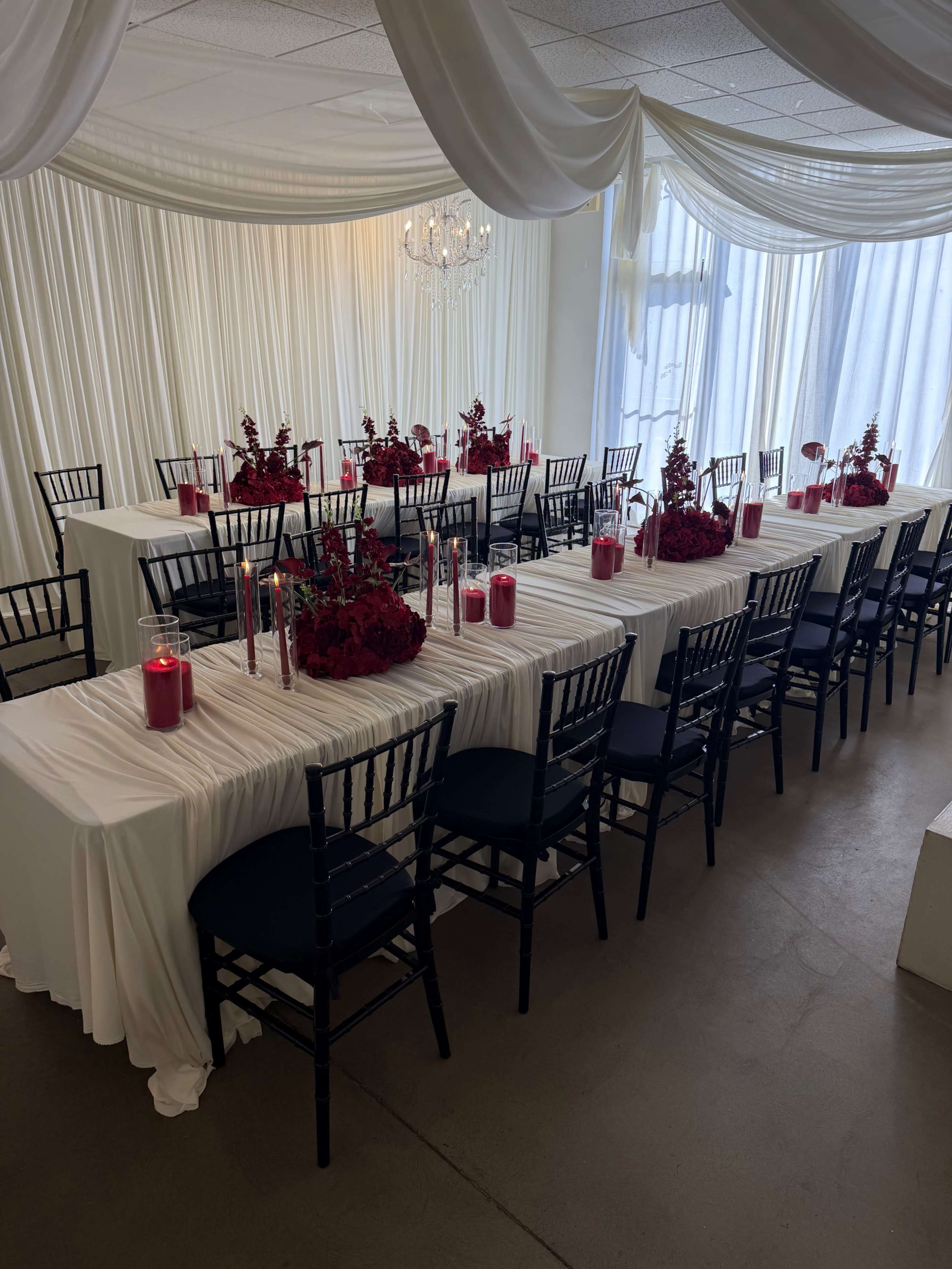 A formal dining setup with long tables covered in white cloth and red floral centerpieces, surrounded by black chairs, is illuminated by candlelight.
