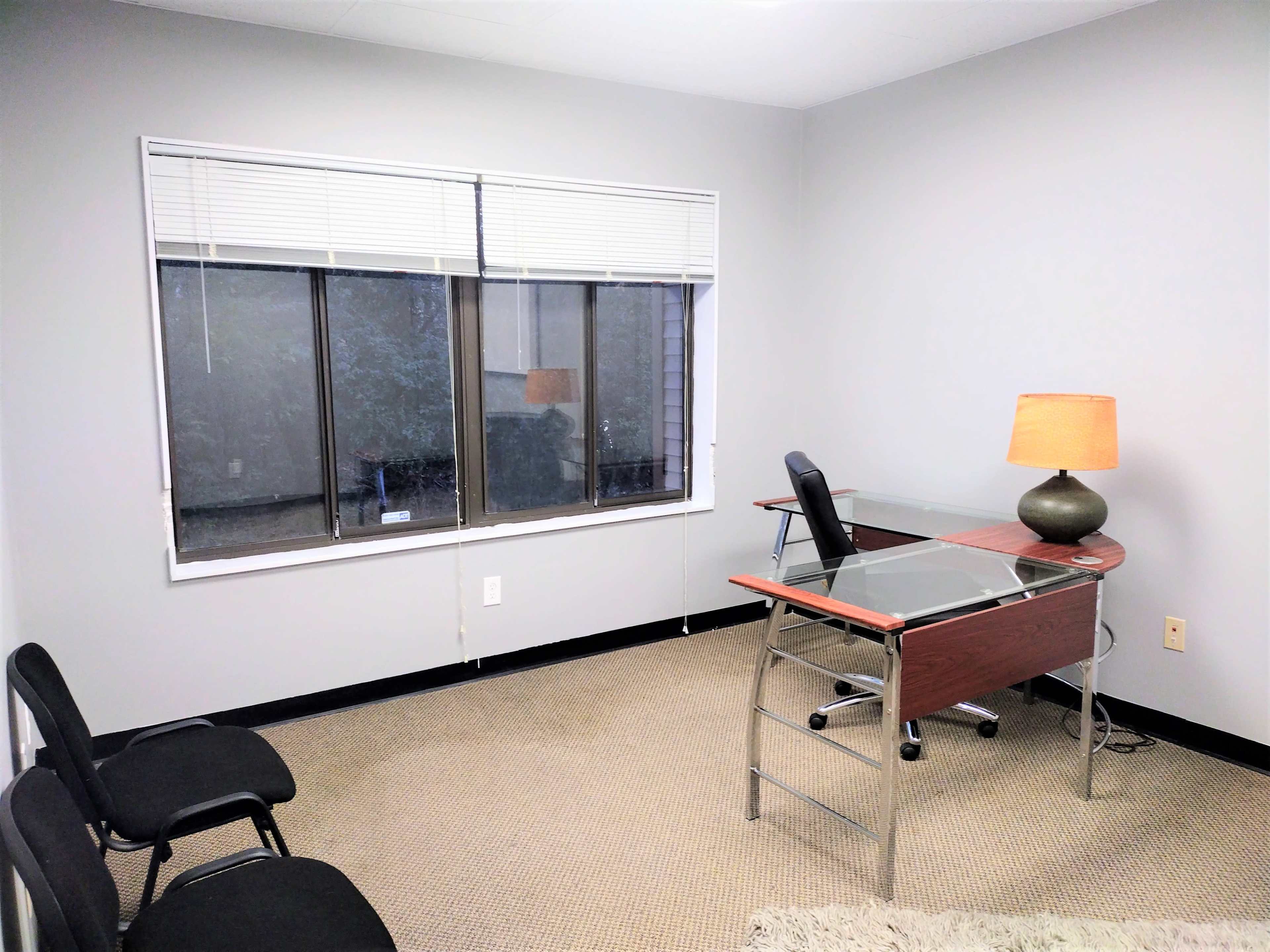 A bare office space with a glass-topped desk, an office chair, a lamp on the desk, and two black chairs against the wall.