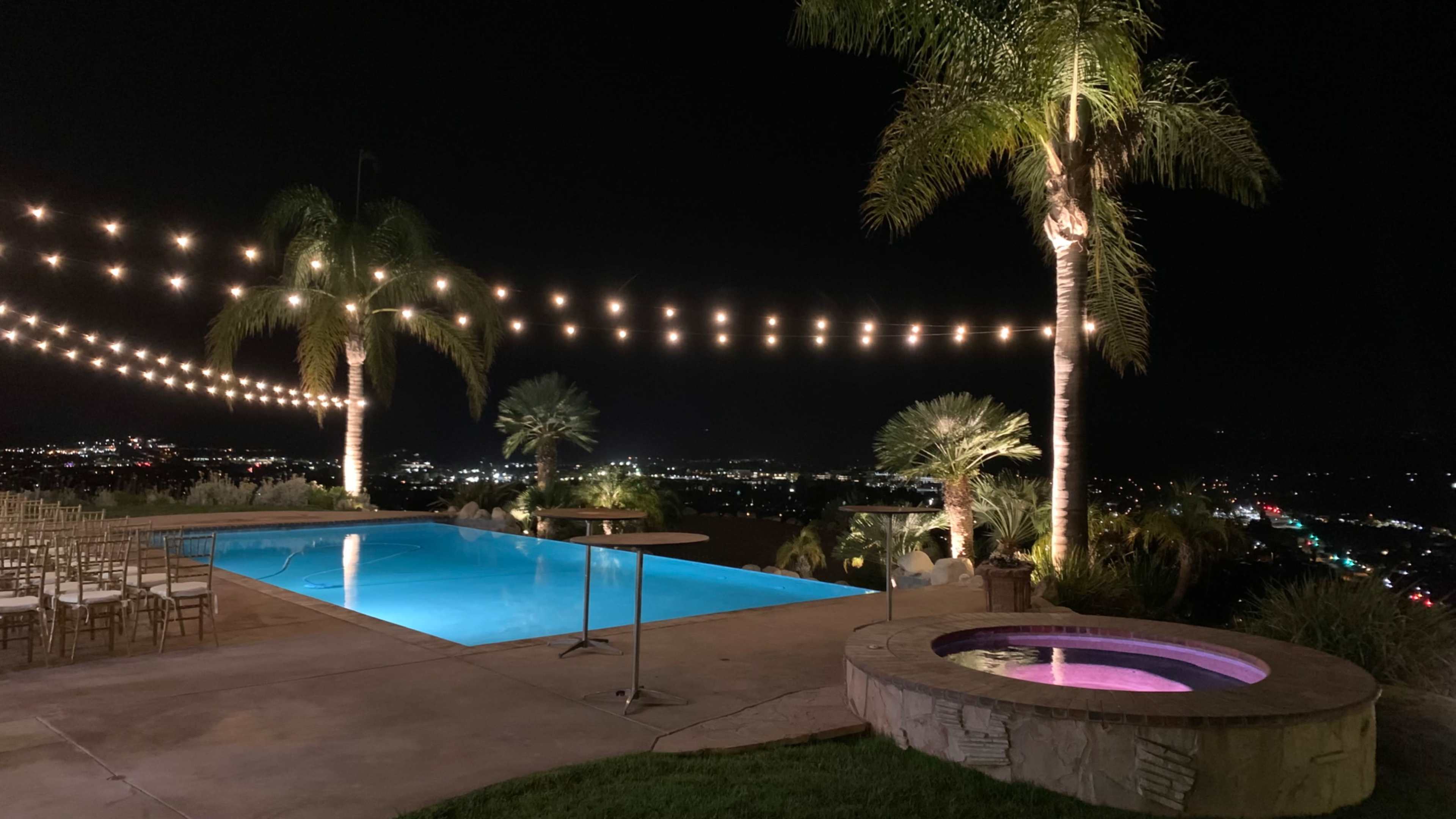The image shows a well-lit outdoor pool area at night, complete with string lights overhead, a hot tub, and palm trees, overlooking a cityscape.