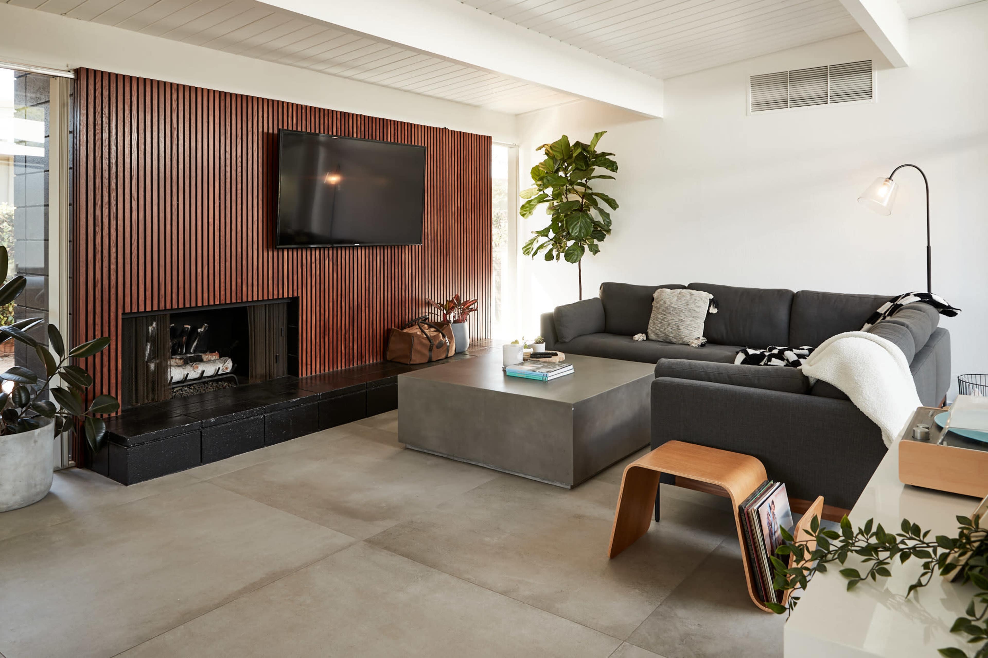 The living room features a dark gray sectional sofa facing a mounted television above a textured wood accent wall, with a concrete coffee table and plants enhancing the space.