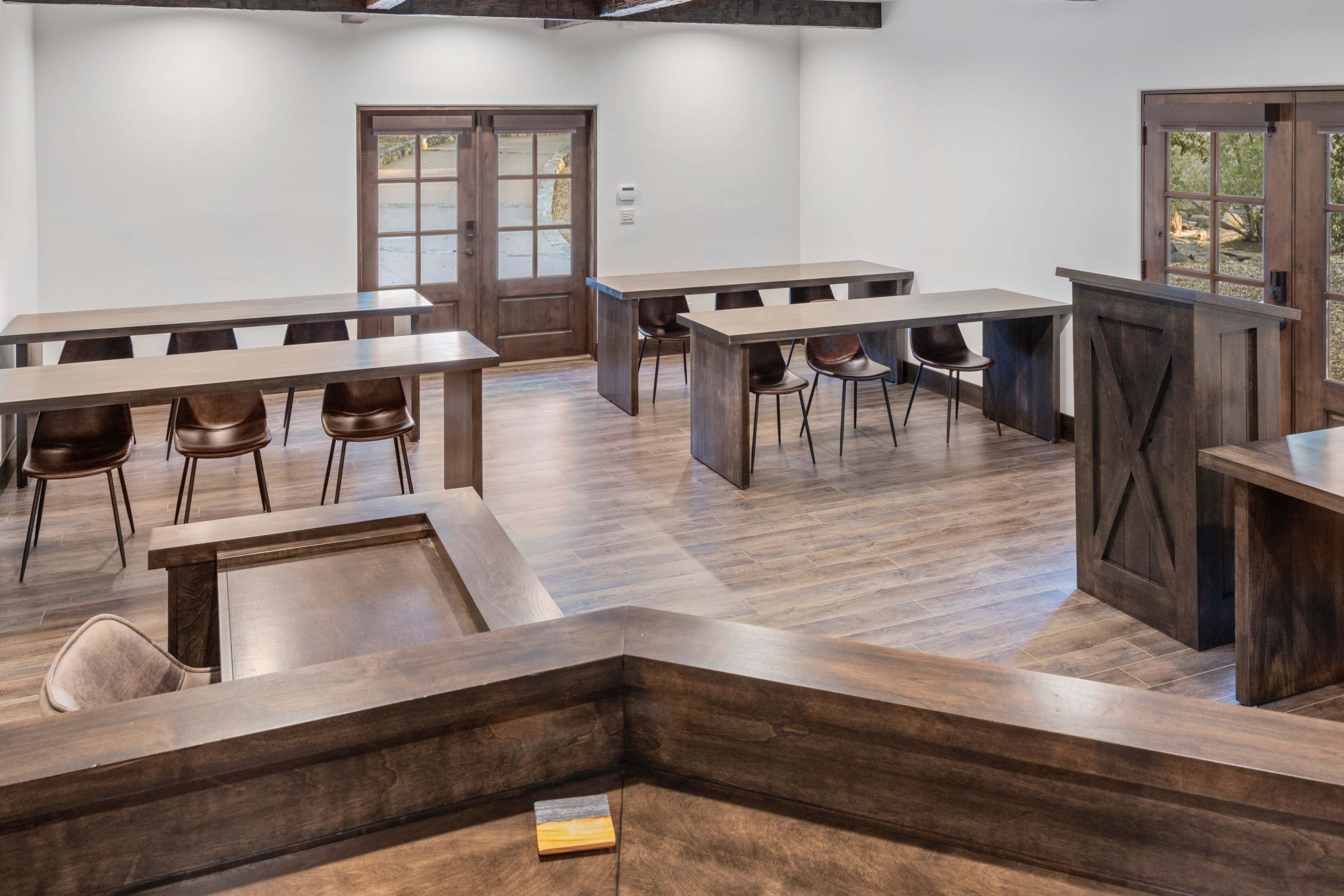 The image shows a spacious, well-lit room with wooden tables and chairs arranged in a classroom-style layout, featuring a large door and windows along one wall.