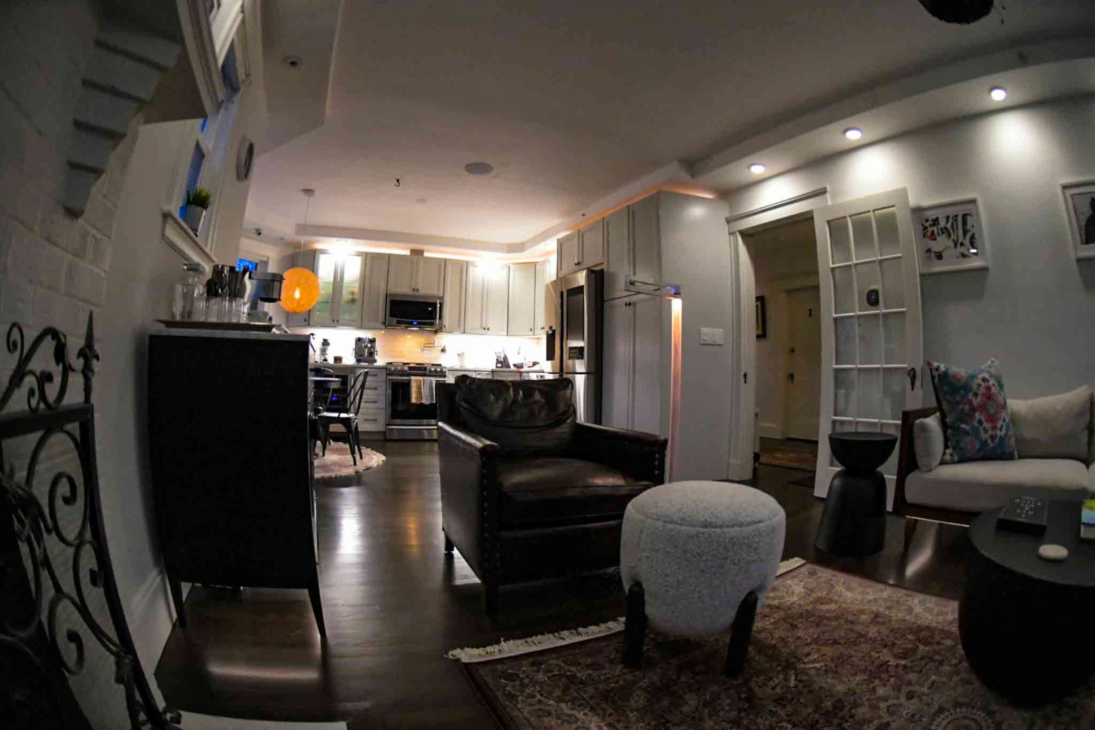The image shows a modern living space with an open kitchen area and a seating area featuring a dark leather chair and a light-colored ottoman.