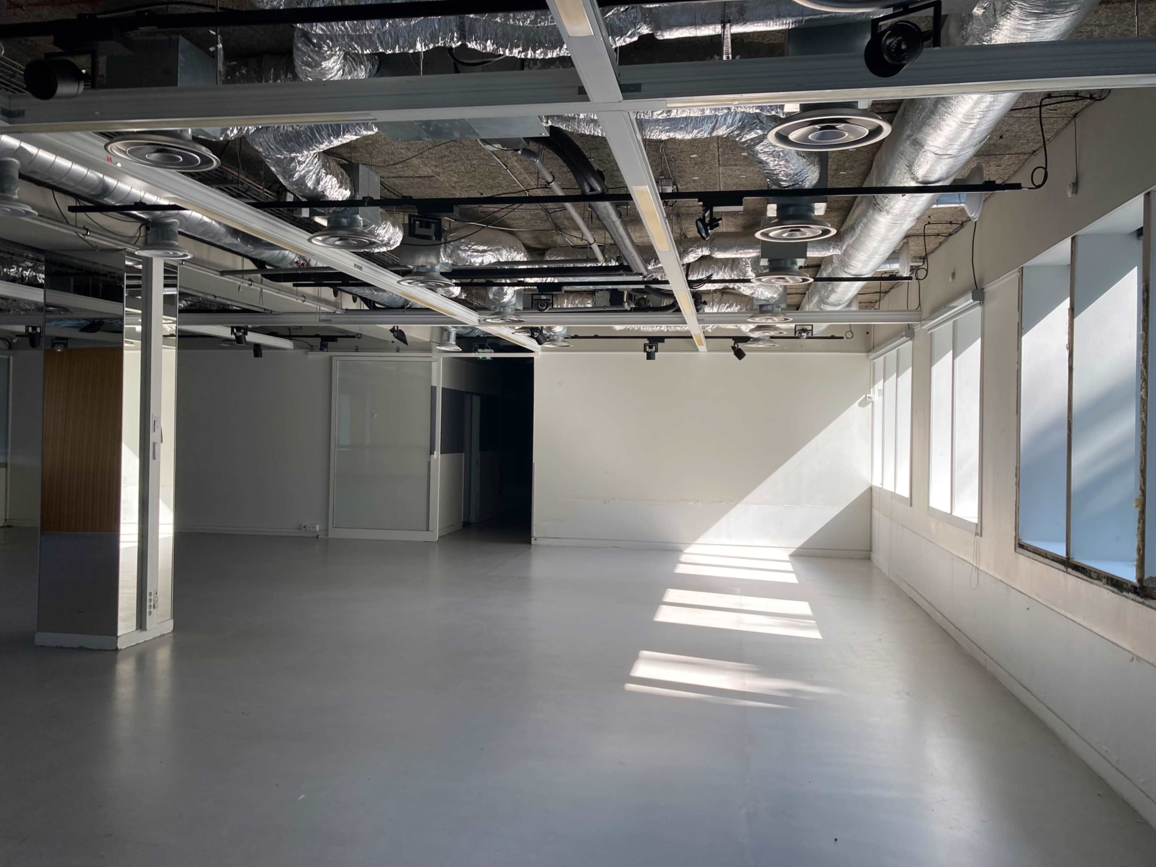 The image shows an empty office space with exposed ceiling ducts and large windows allowing natural light to enter.