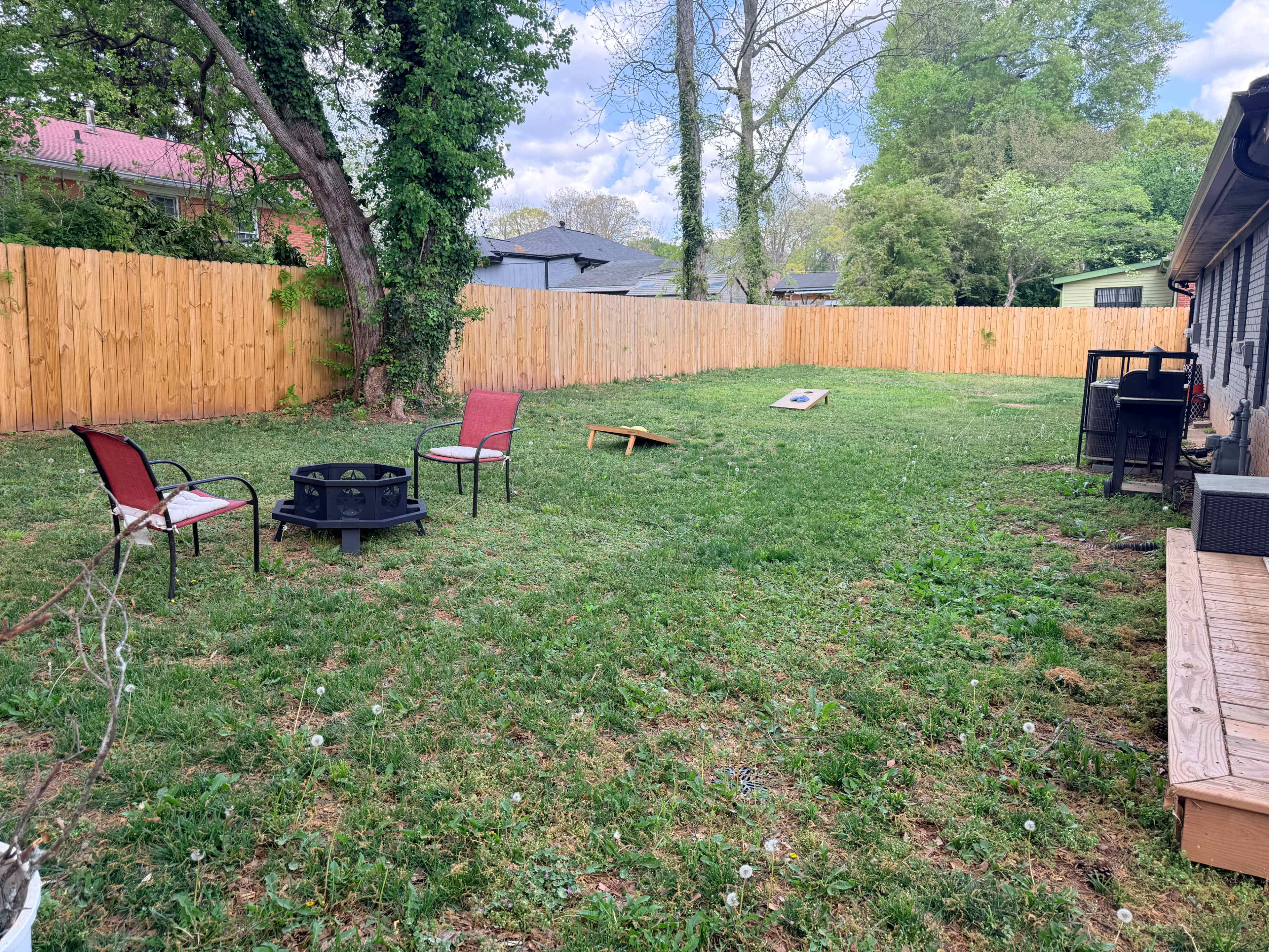 The image shows a grassy backyard with a fire pit surrounded by two chairs, a cornhole game set up, and a wooden deck adjacent to a brick house.