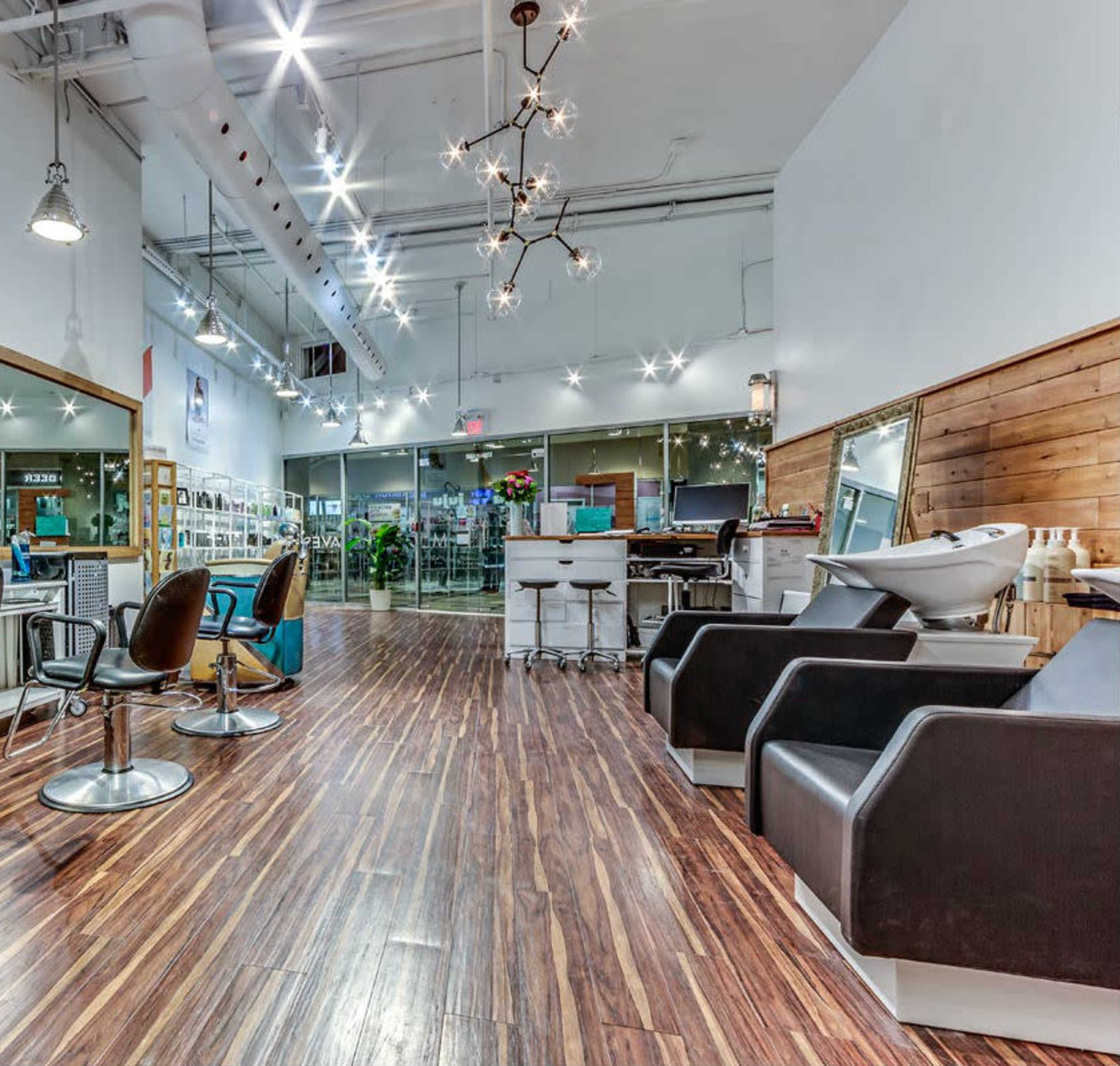 A modern hair salon with sleek wooden flooring, styling chairs, and a large mirror, illuminated by bright overhead lights.